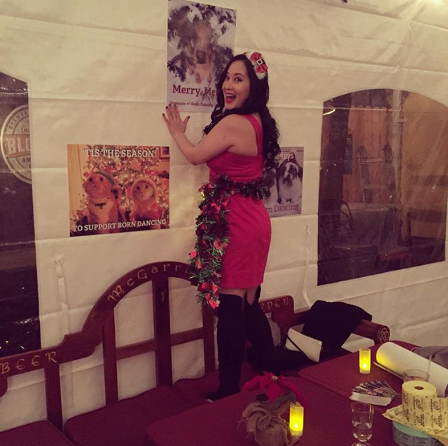 Board member Jessica Gallagher in a red dress and Christmas garland standing on a bench to hang a Born Dancing poster at the Holiday Party.