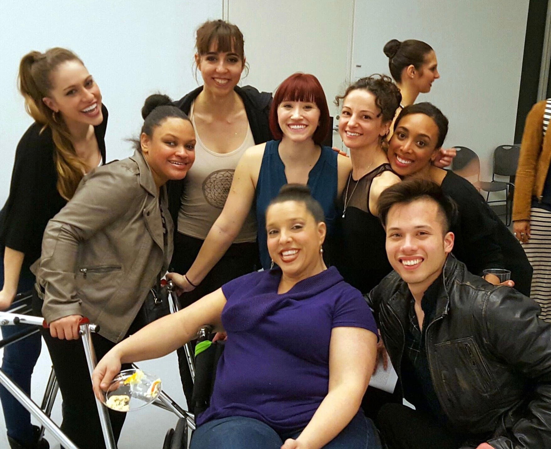 Seven female dancers and one male dancer in street clothes clothes smiling for group photo. Some are seated or standing using wheelchairs or walkers