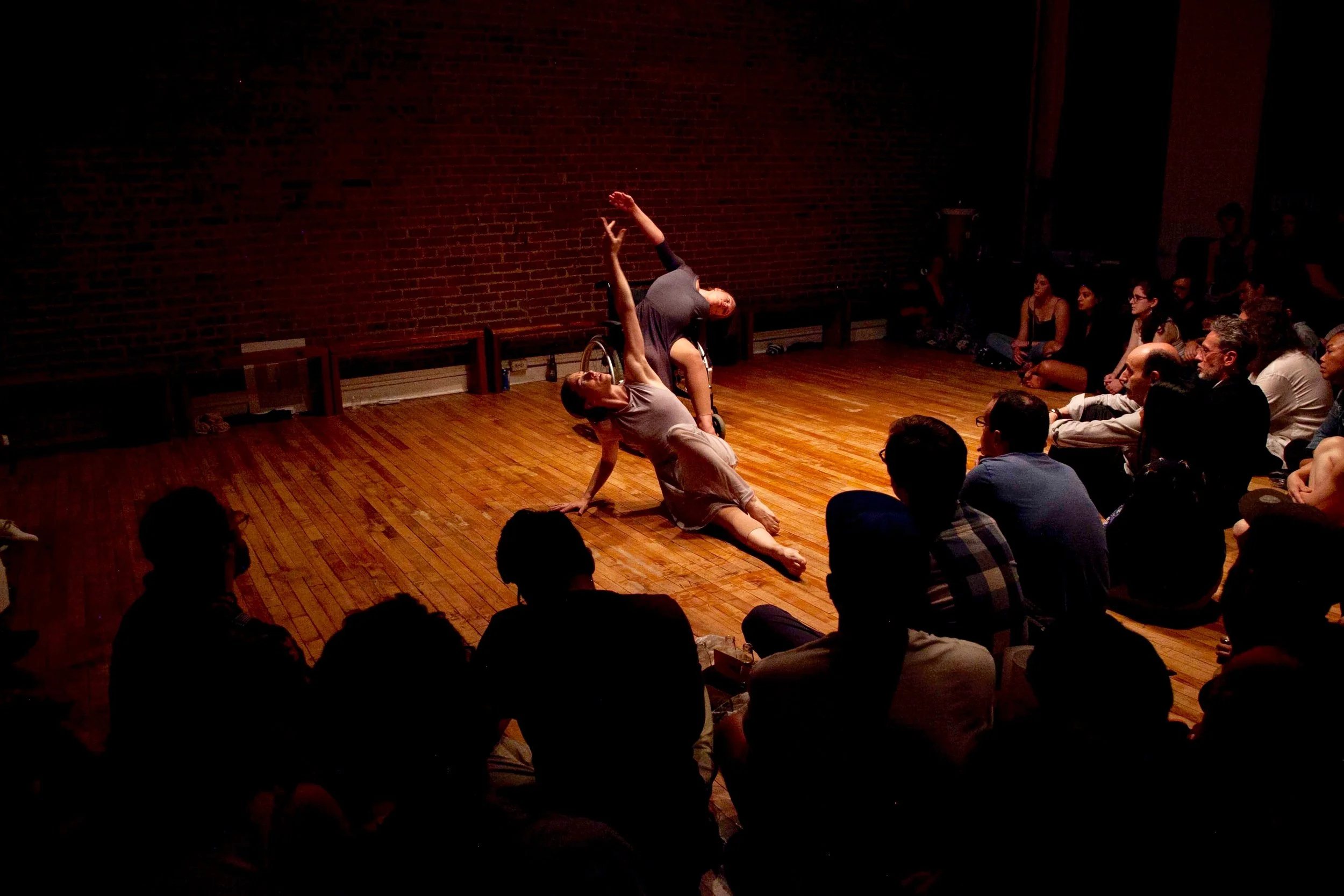 Madeline Charles and Madison Ferris. (Photo: Kyle Netzeband). (Audience seated on floor around two female dancers in grey dresses reaching up high. One woman is seated on the floor, the other in a wheelchair).