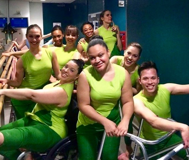 Nine dancers in bright green costumes posing for the camera backstage and smiling.