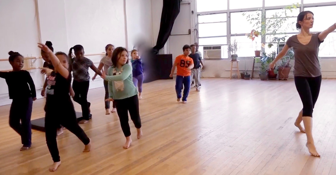 Choreographer Melissa and nine student-dancers rehearsal, arms outstretched in front while walking.