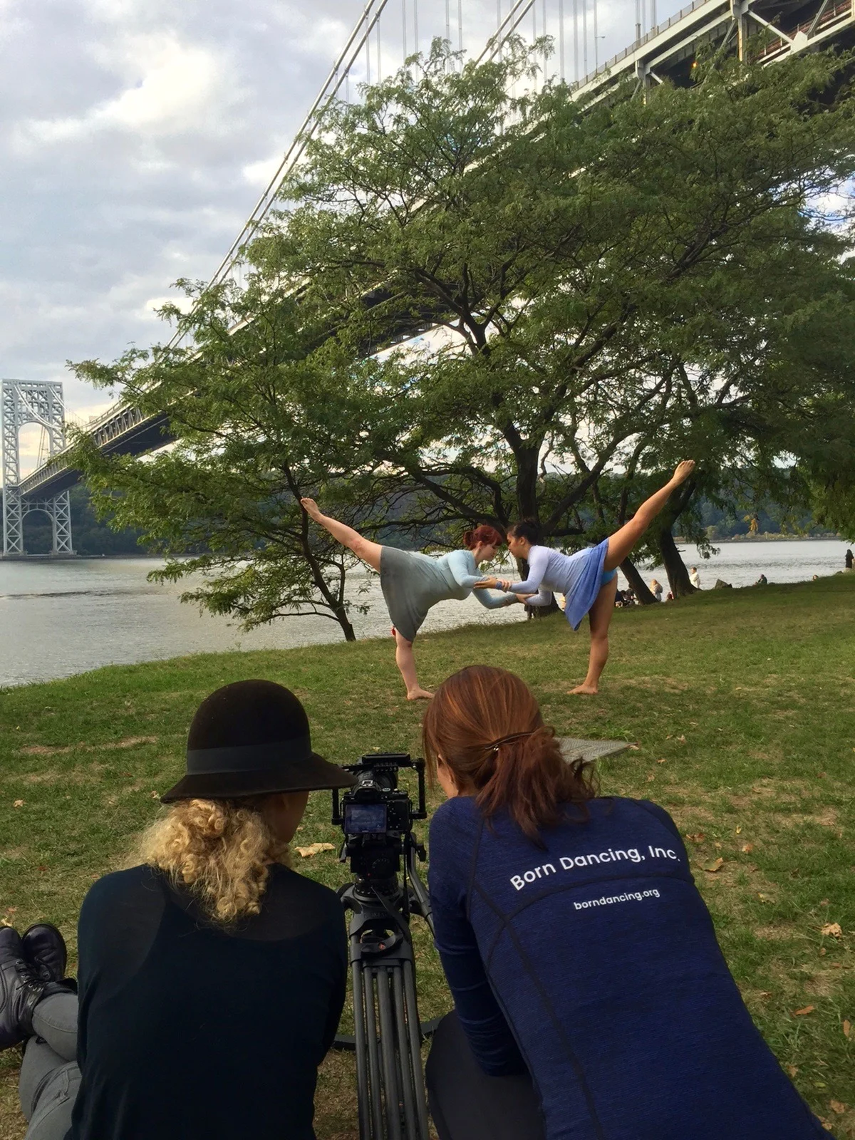 Choreographer Melissa and Videographer Cheyenne seated at the camera filming two female dancers in arabesque on a grass field underneath the George Washington Bridge.