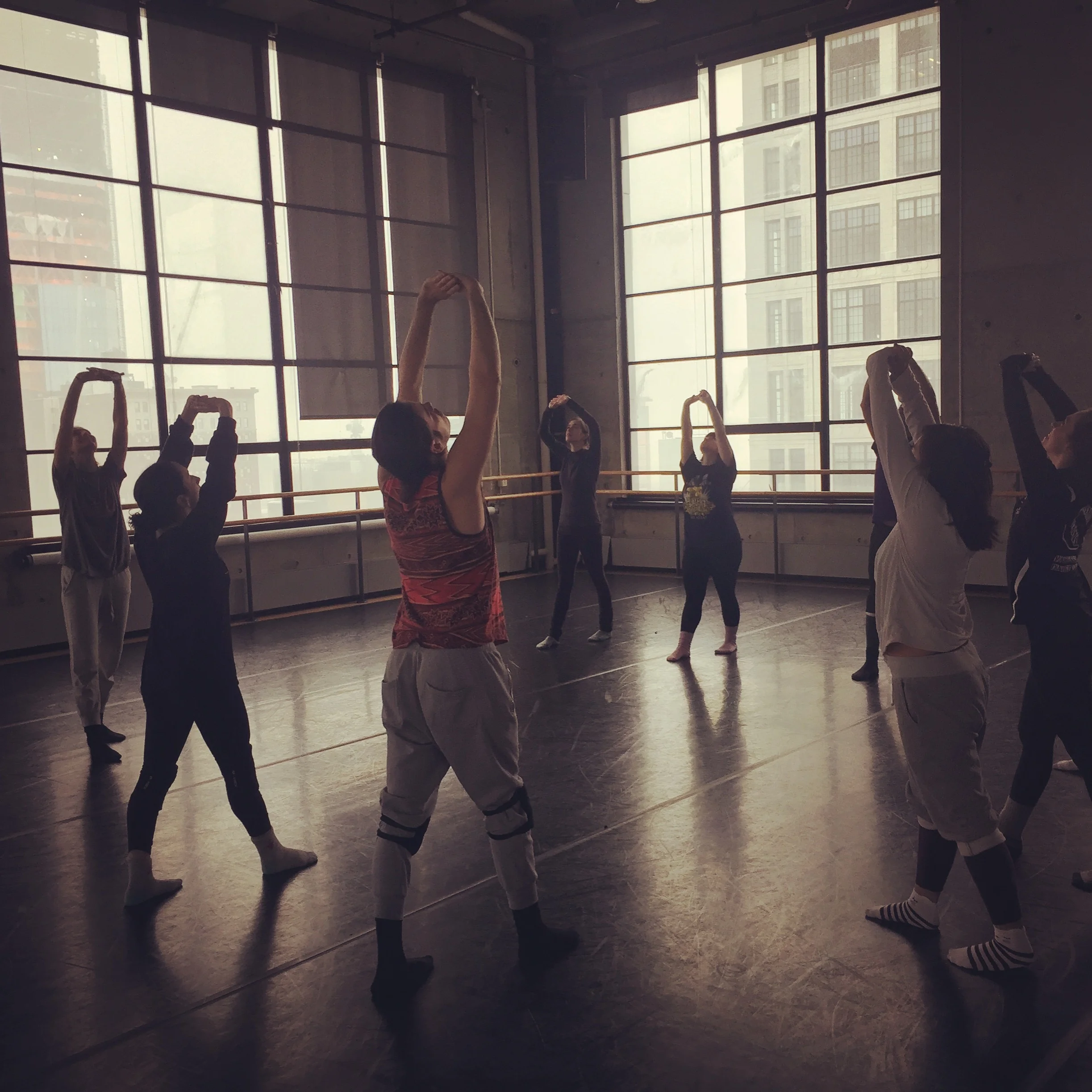 Dancers standing in a square formation with their arms up high and hands flexed and touching in a dance studio with huge windows.