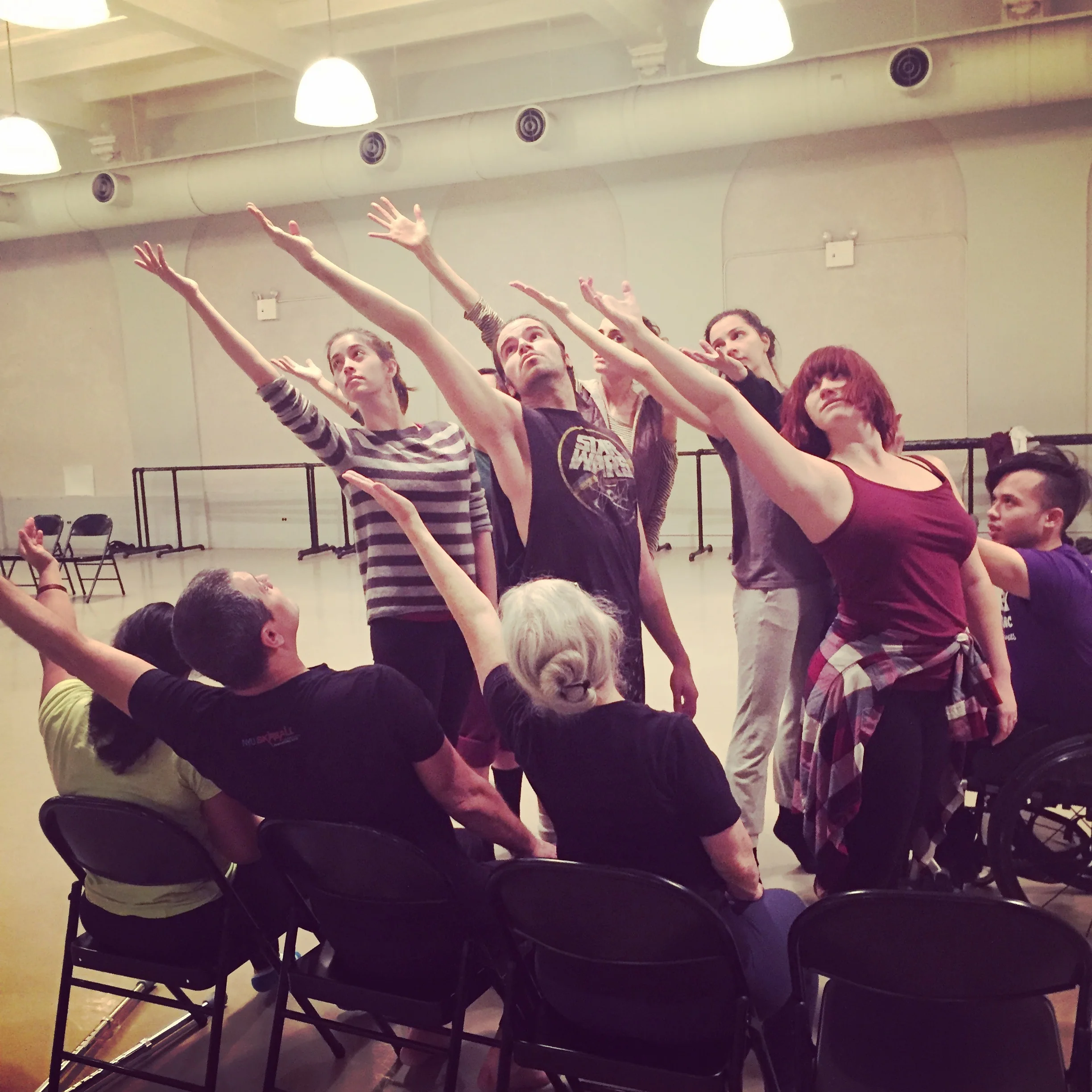 Dancers seated on chairs and standing in a small group in rehearsal with one arm extended behind them and reaching up.