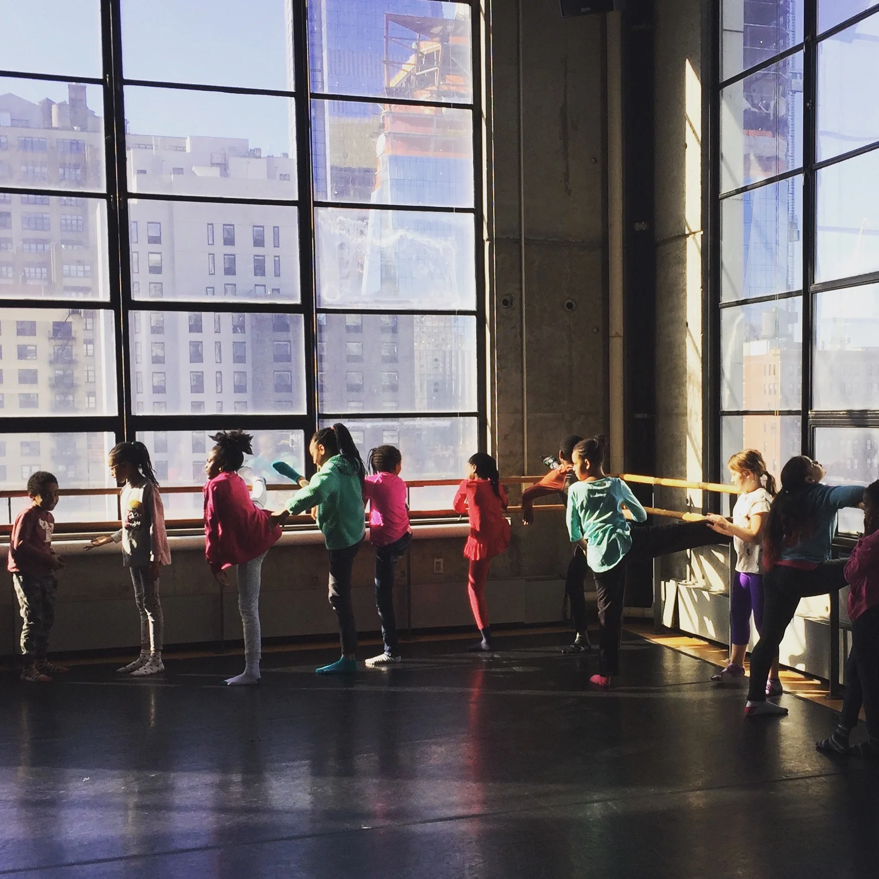 4th and 5th grade student dancers standing and stretching their legs at the barre in a large dance studio with big windows looking out on neighboring buildings.