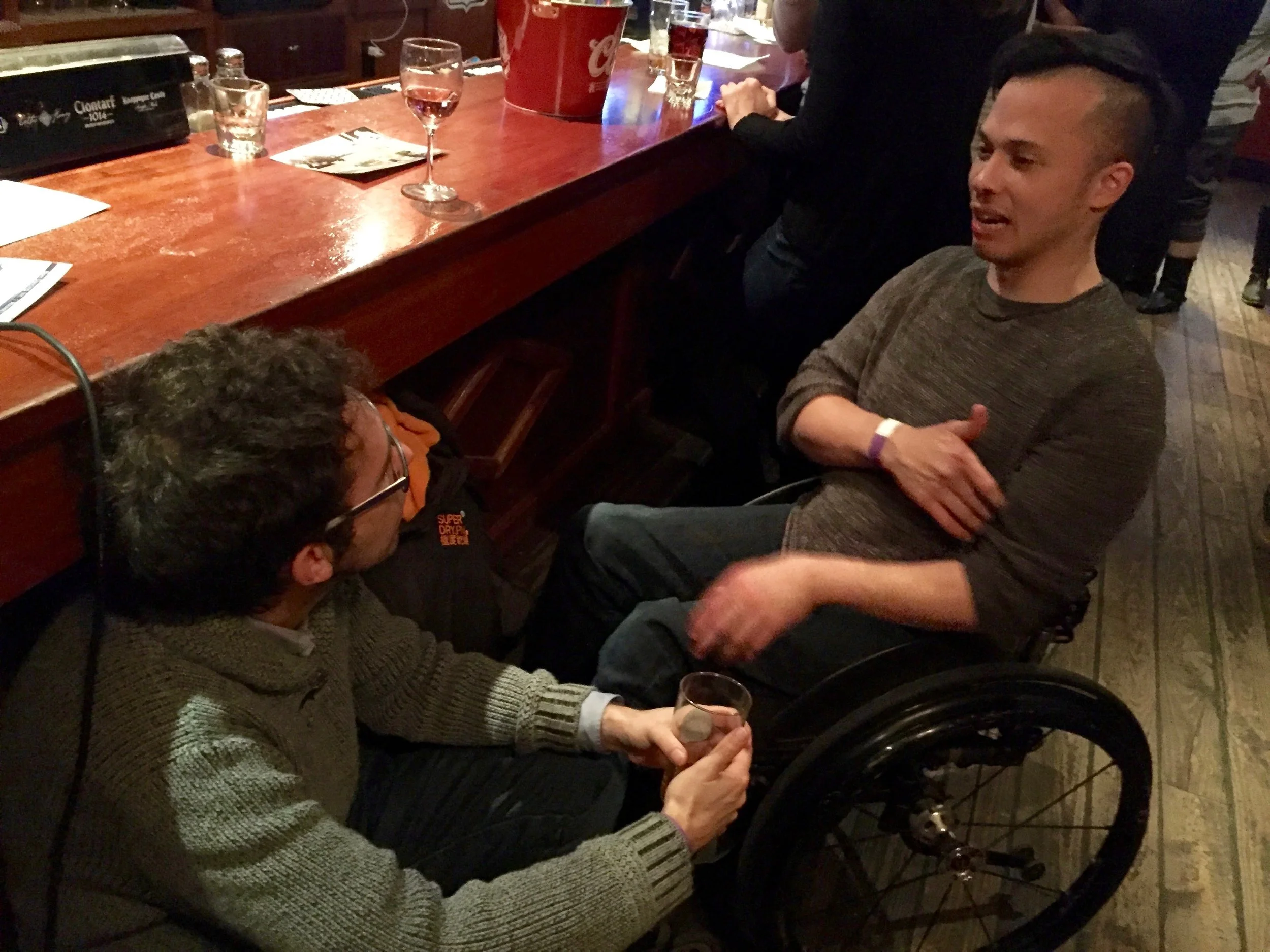 A man and a male dancer using a wheelchair in conversation at a bar at our Holiday Party.
