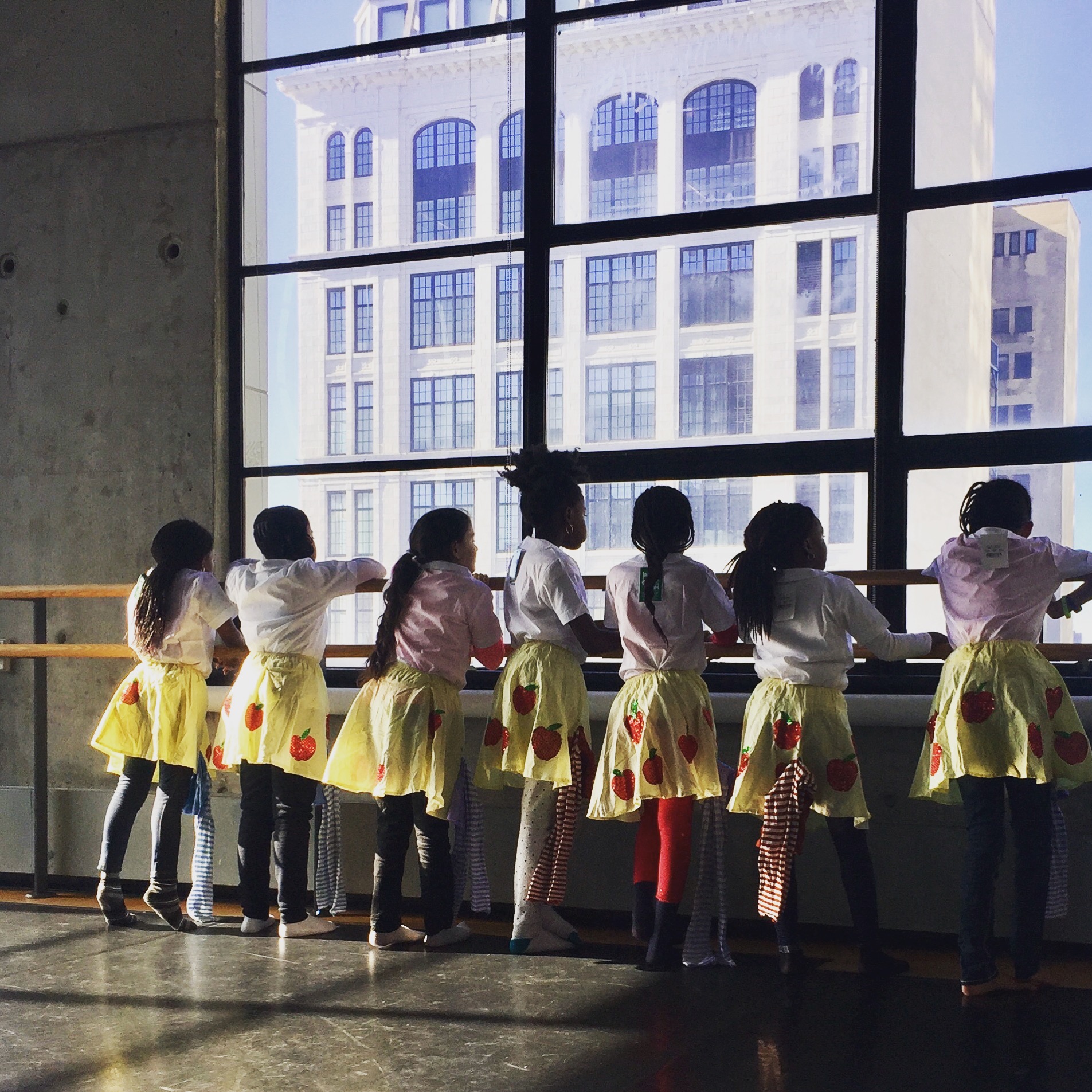 Seven girls in a mix of dance attire and costumes leaning on the barre while looking out the window of a dance studio.
