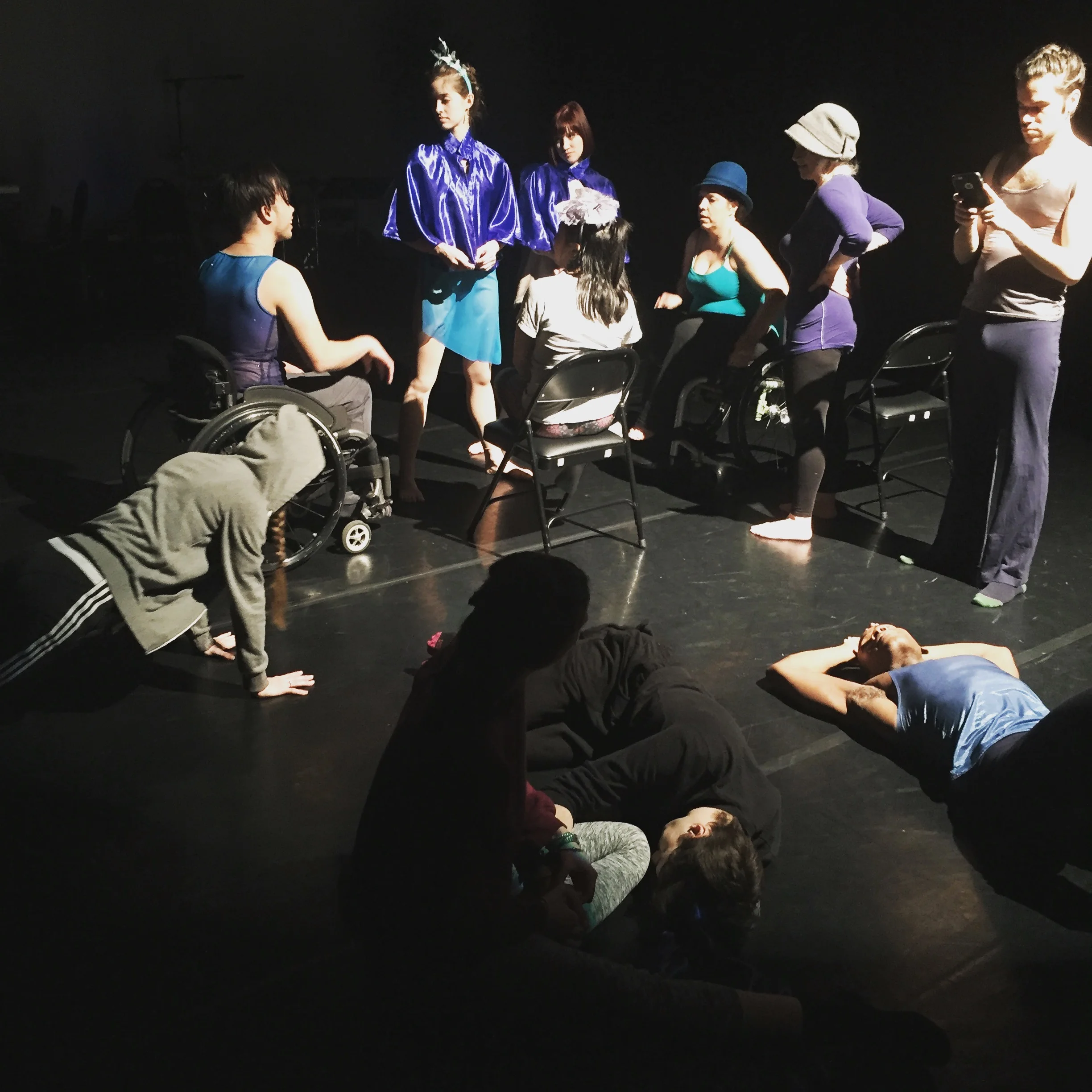 Nine dancers sitting and standing around in dance attire and partial costumes while waiting during tech rehearsal at the theater.