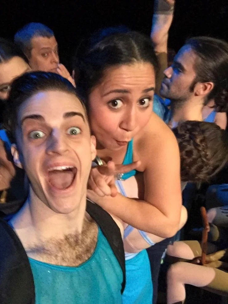 Dancers dressed in blue and teal costumes taking a selfie while dancing on stage portraying a scene on a NYC subway.