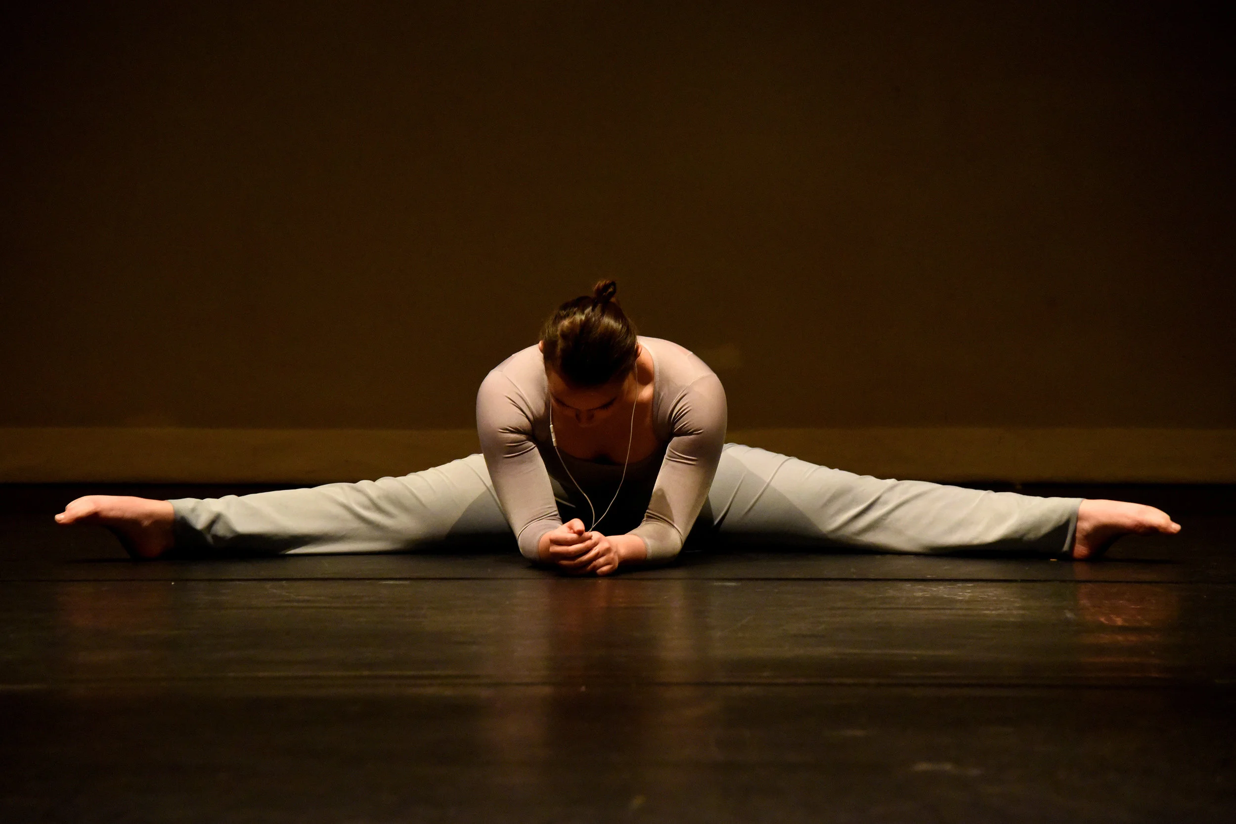 Dancer Katie Mollison warming up in costume stretching in a large straddle on a dimly lit stage with earphones in.