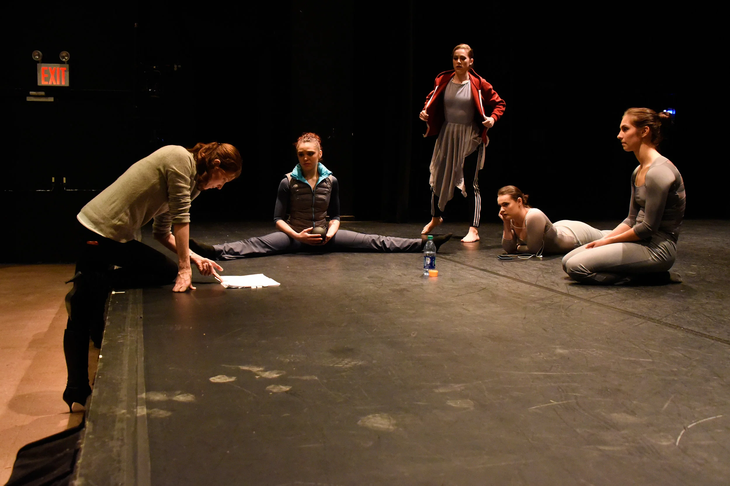 Choreographer Melissa looking at her notes from the side of the stage while four female dancers wait and stretch on stage.