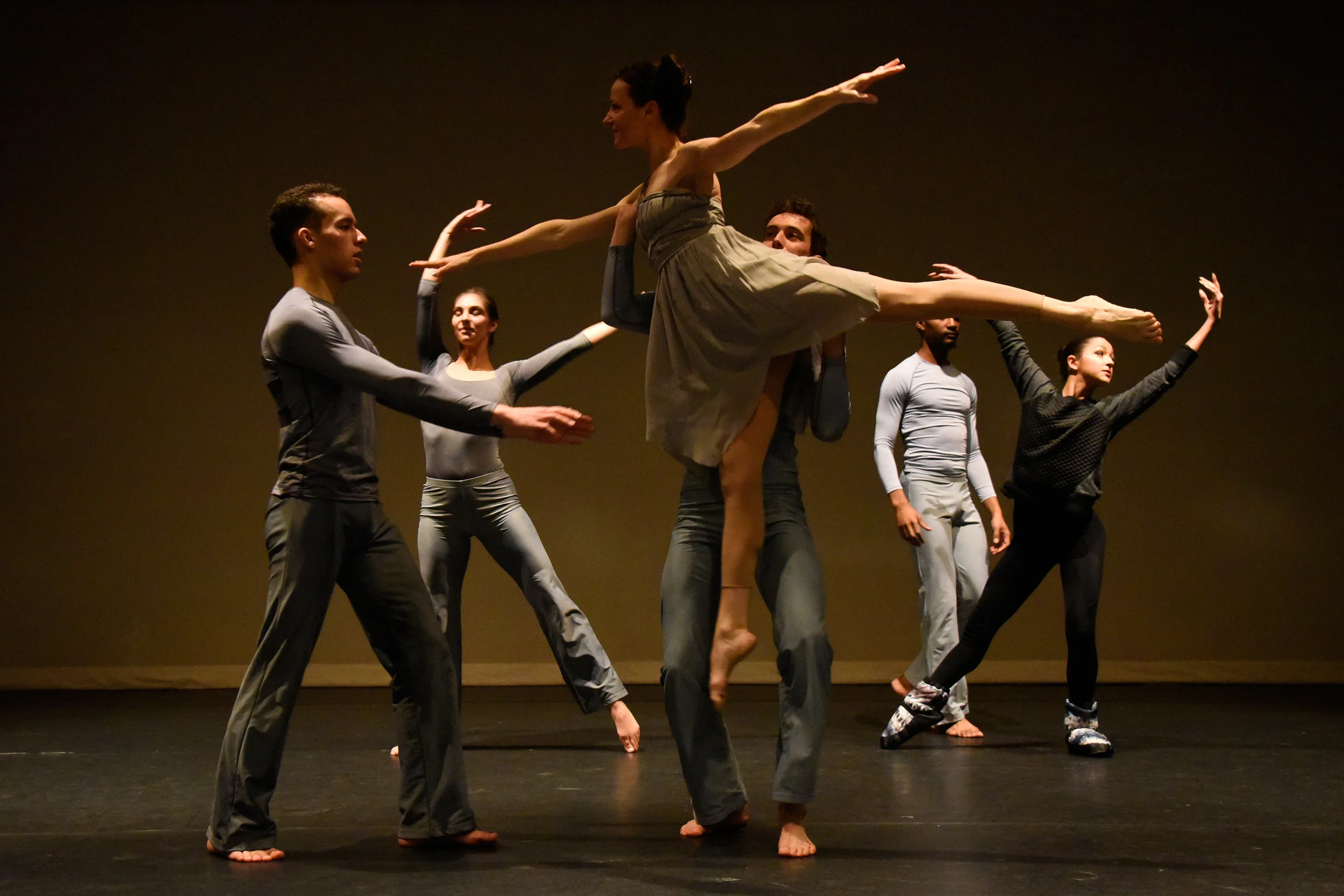 Dancer Nicole Speletic gets lifted while her leg is behind her in arabesque and her arms reach out and she is passed to a different male dancer. Three other dancers warm up nearby on stage.