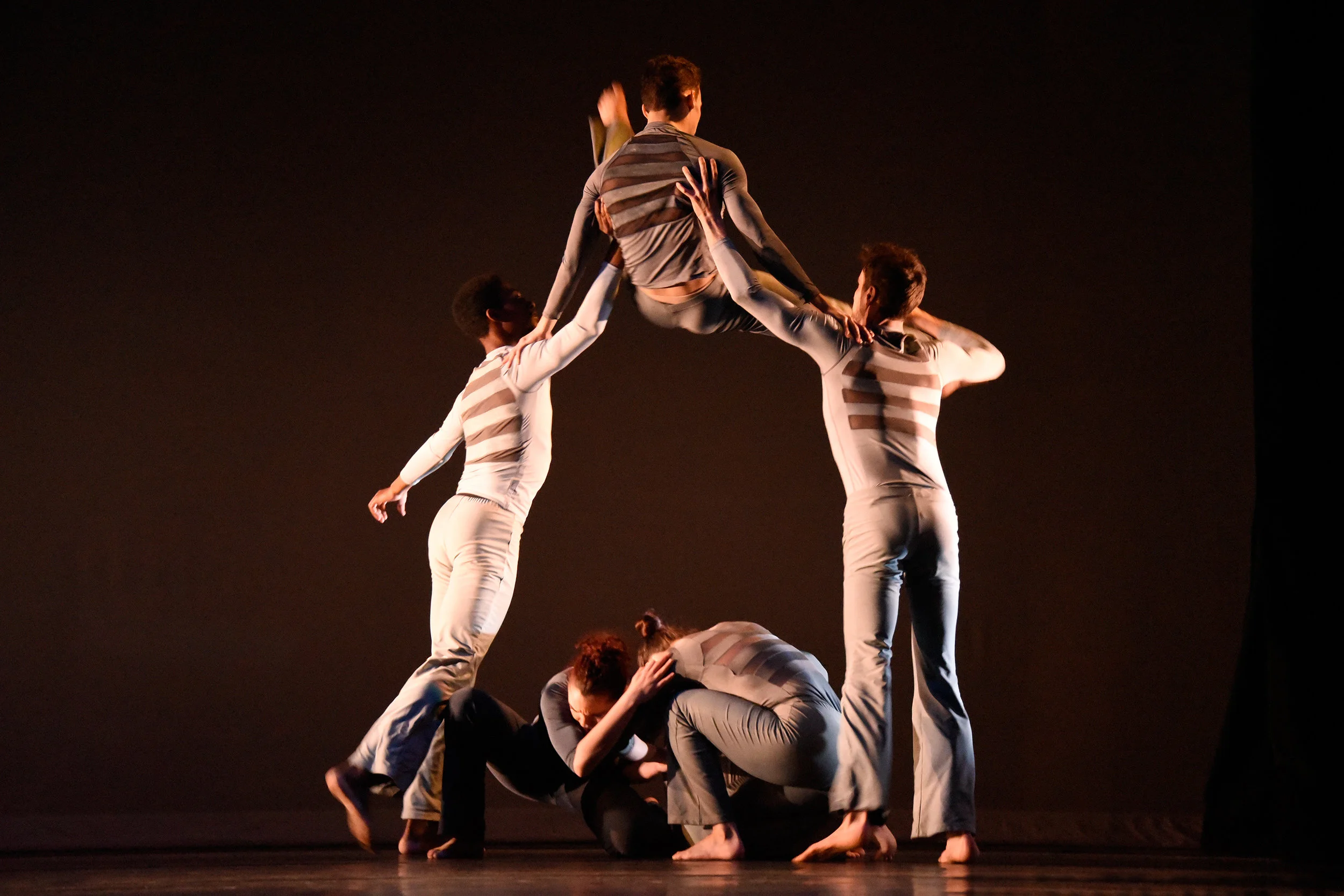 Grey. Male dancer jumps over two women huddled together low on the floor by pushing off with his hands against the shoulders of two men standing on either side of the women.