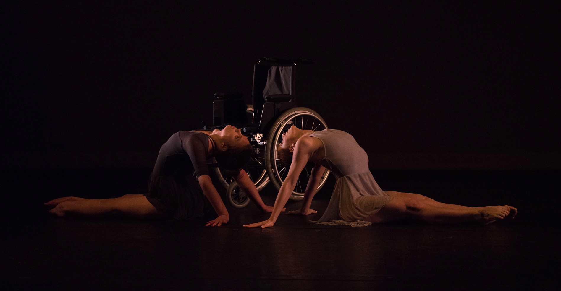Grey. Madison Ferris and Madeline Charles are seated back to back in front of a wheelchair and arch their backs towards each other. Both are wearing light and dark grey leotards and skirts.