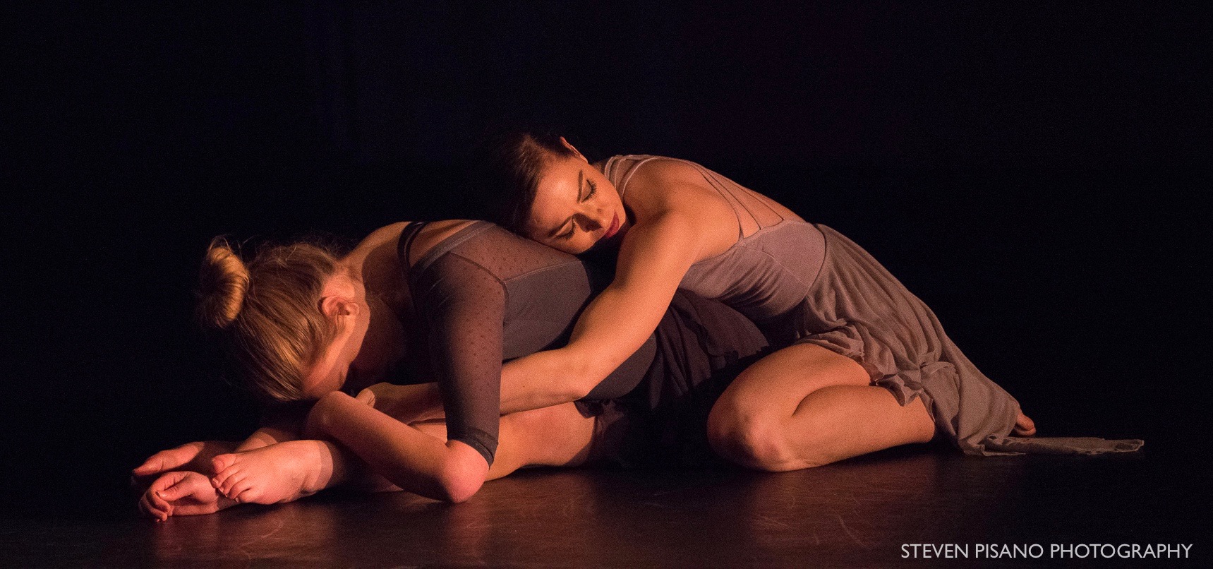Grey. Madison Ferris and Madeline Charles are seated behind each other on the floor as they fold forward and hold each other.