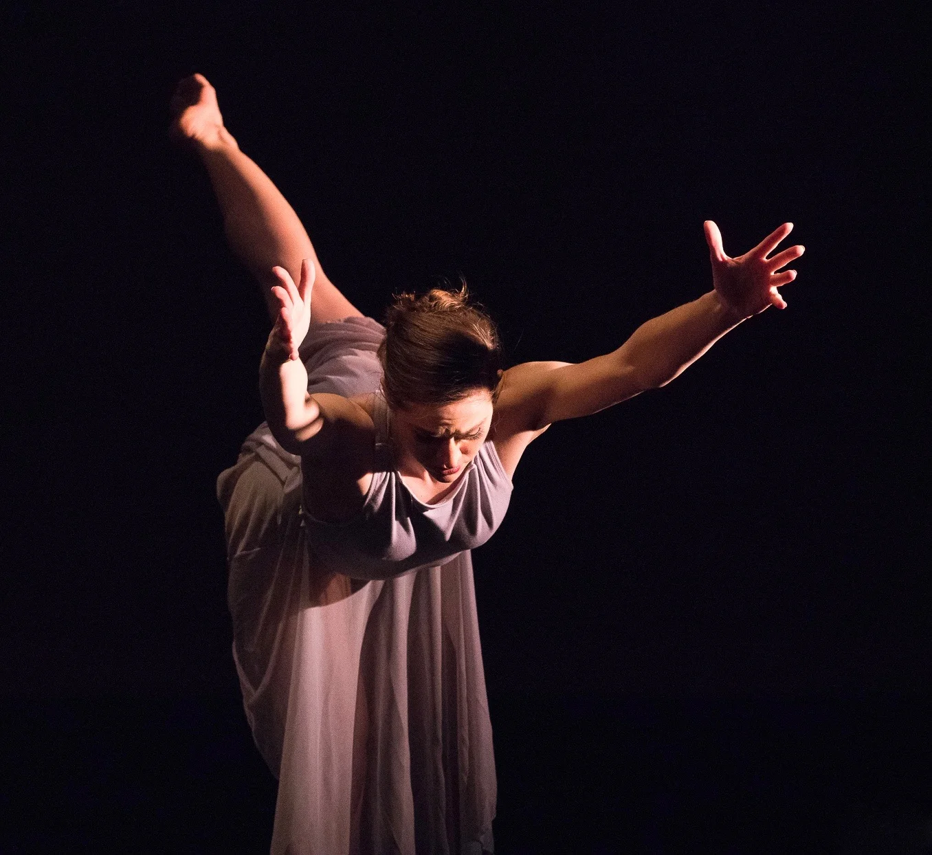 Grey. Madeline Charles stands on her right leg and has her left leg extended high behind her in arabesque as she reaches both arms forward and up.