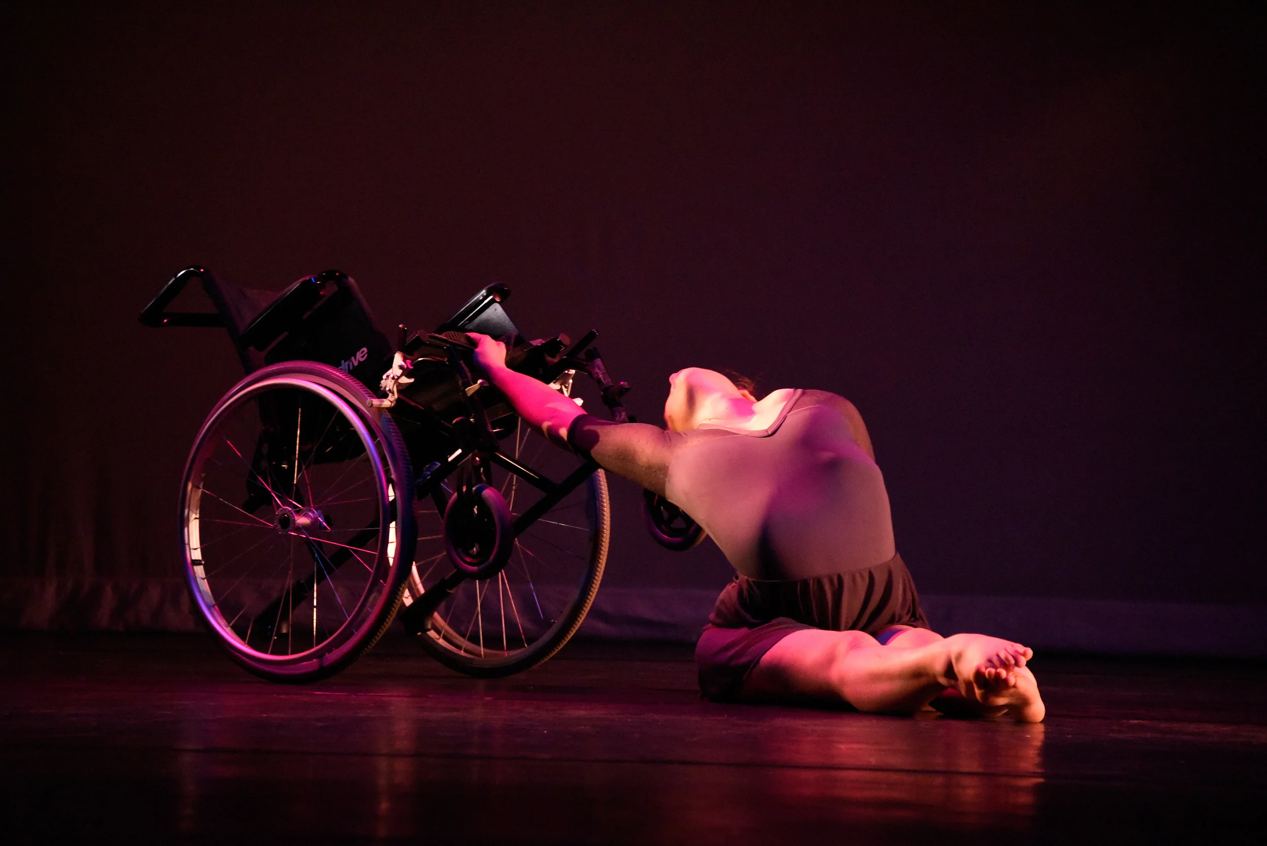 Grey. Madison Ferris sits on the floor with her legs straight in front as she reaches back and pushes against the seat of her wheelchair into a big arch.