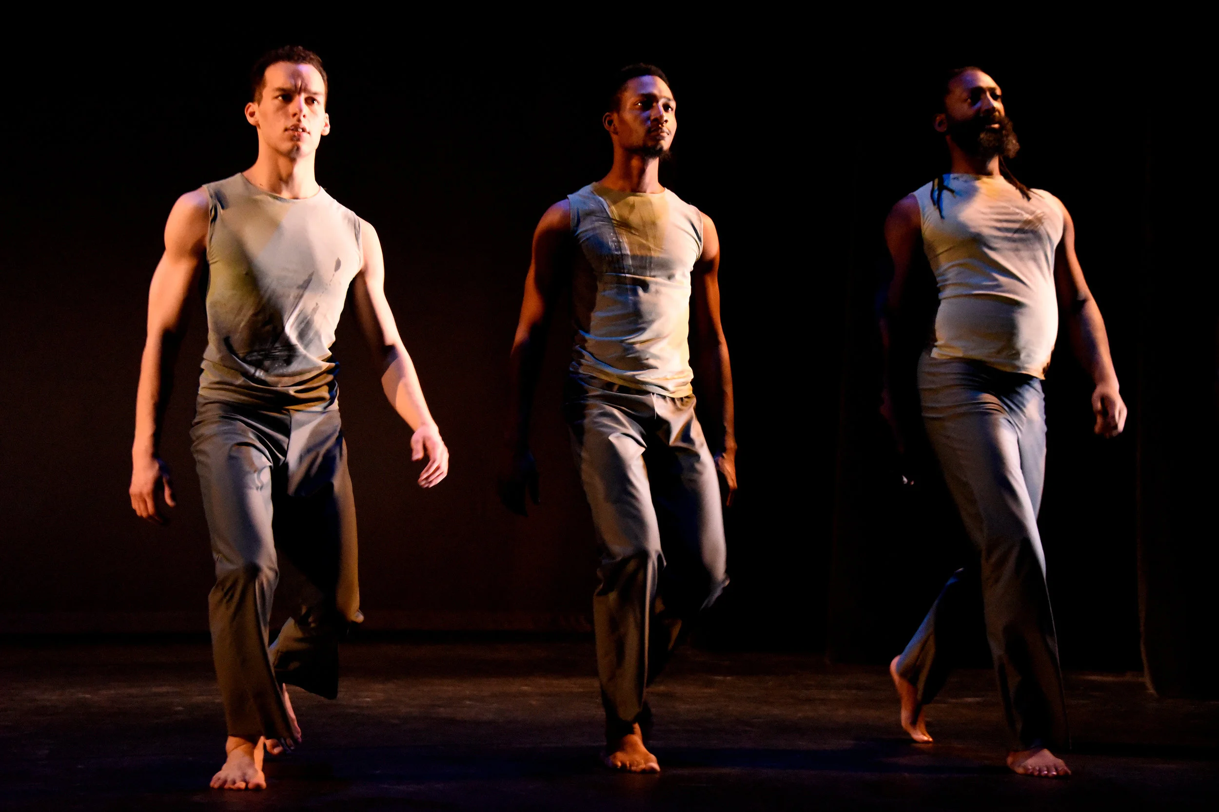Grey. Close up of three male dancers in sleeveless grey costumes walk in a straight line towards the audience.