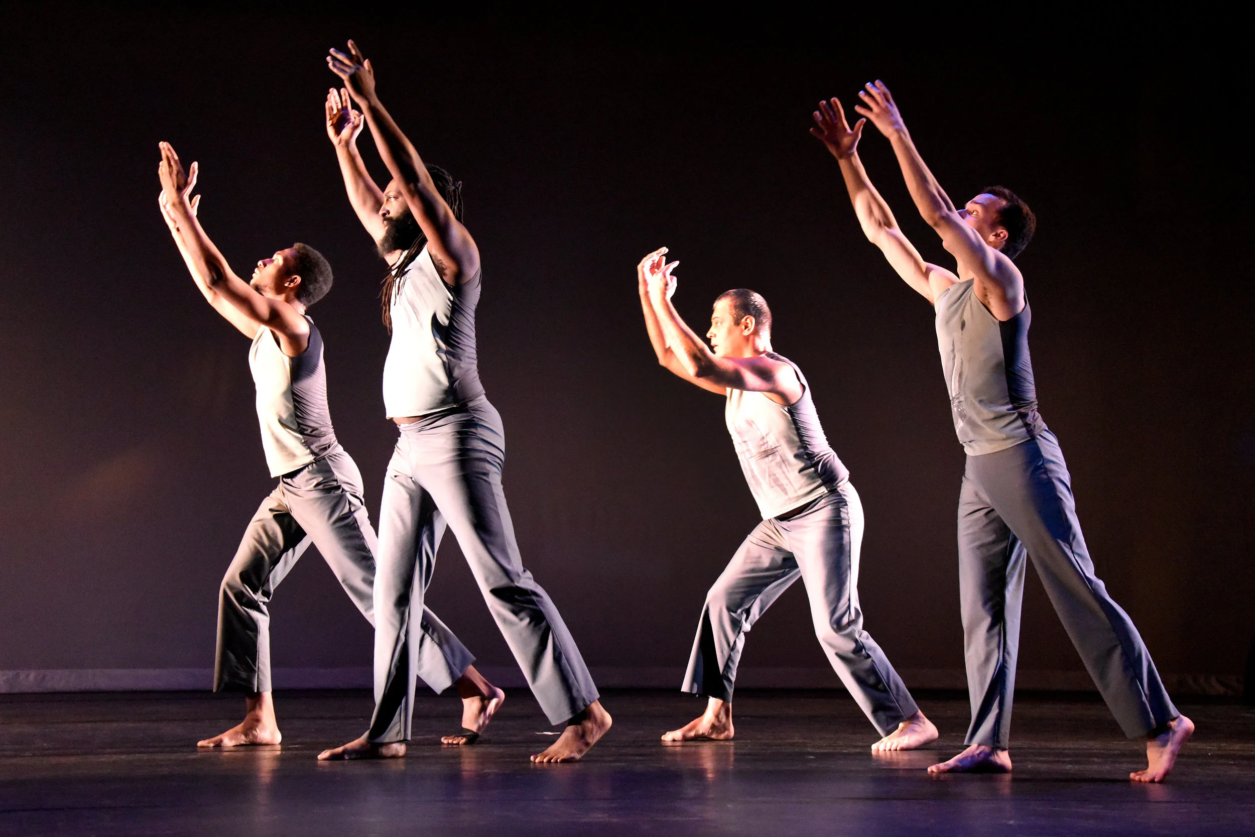 Grey. Four male dancers reach up with both ares as they face the same direction and lunge with their legs.