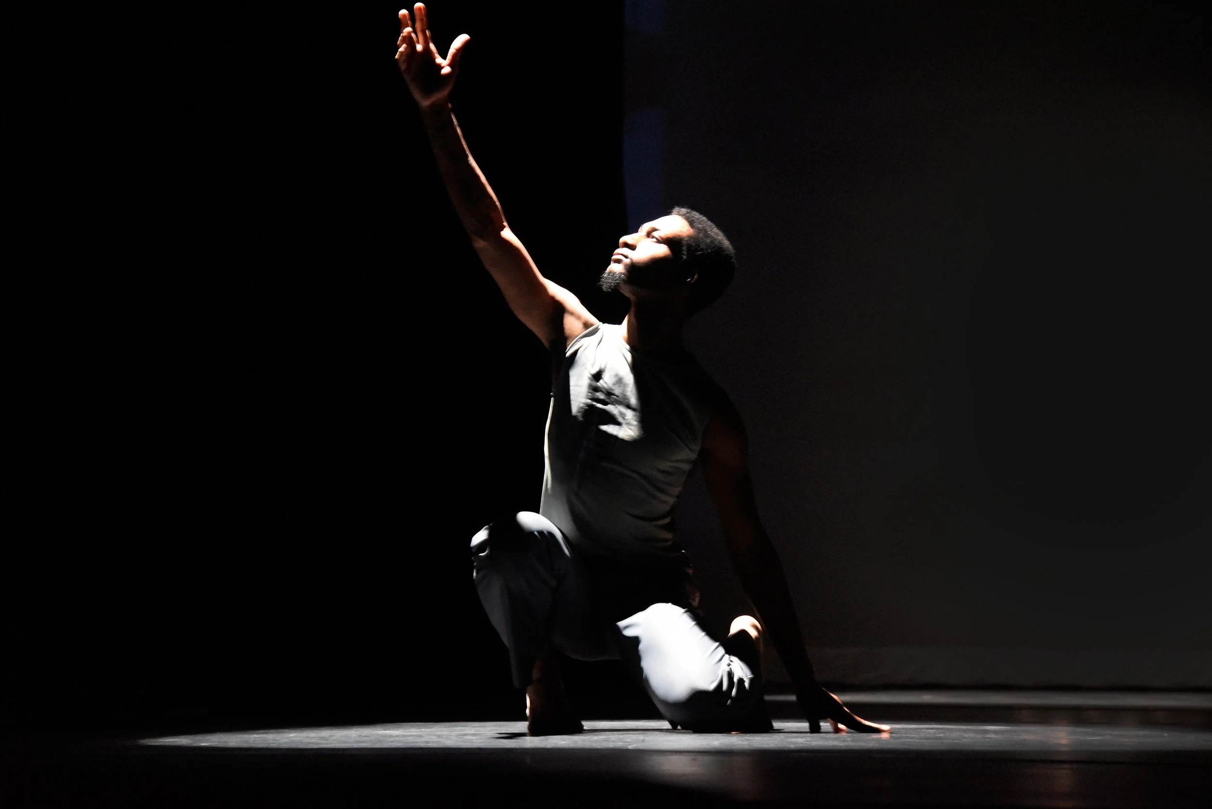 Grey. On a dark stage in a small lit circle dancer Dominique Lockett sits on one knee and solemnly reaches one arm up as he looks up in the same direction.