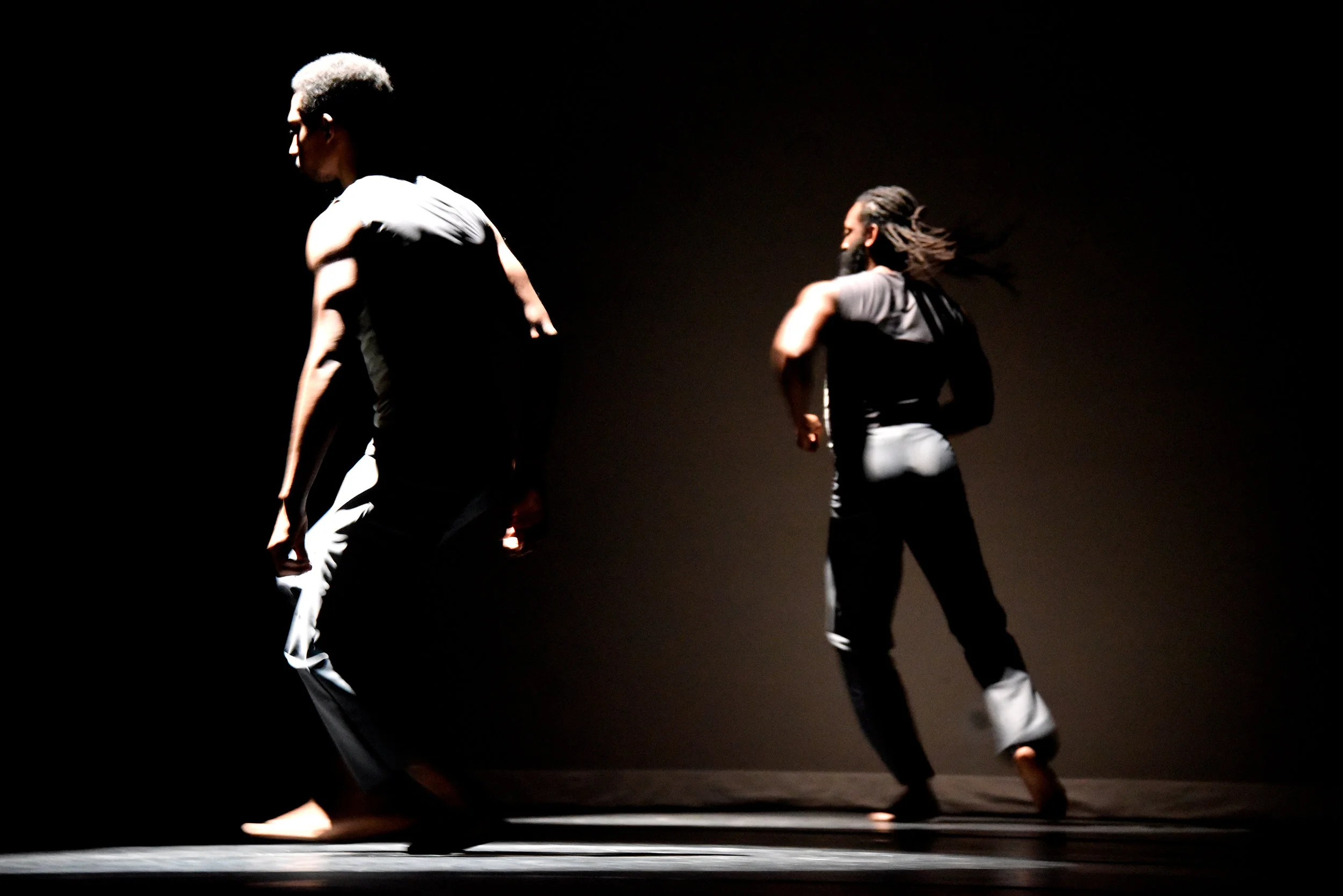 Grey. The stage is dimly lit as dancers Samuel Caraballo and Antoine Hunter start to turn and run.