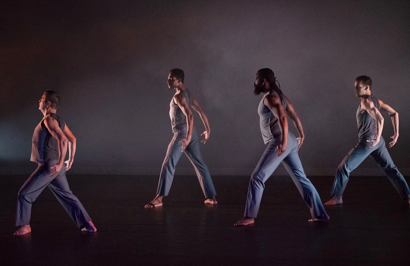 Grey. Four male dancers in sleeveless grey costumes stand in a lunge with bent arms behind them on a dimly lit and foggy stage.