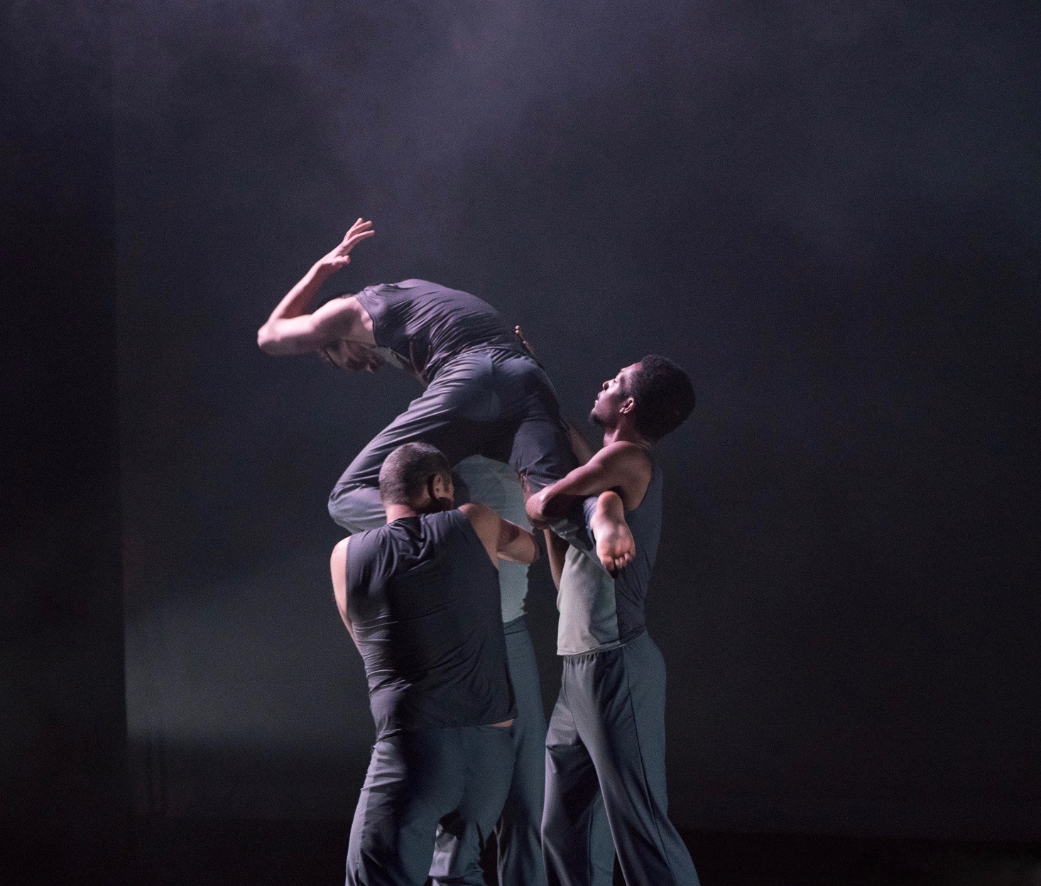 Grey. Male dancer stands on the hands of three other male dancers and is lifted up as he contracts in and looks down.