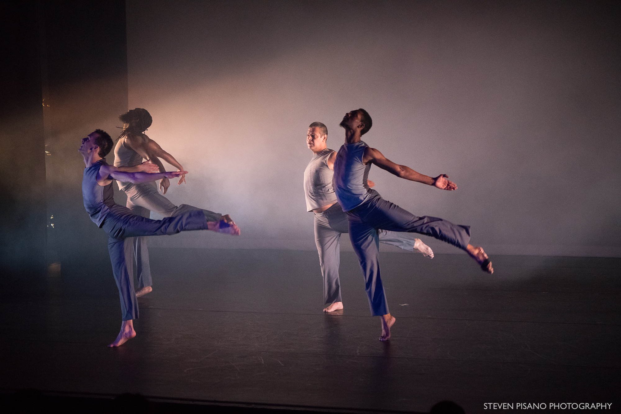 Grey. Four male dancers in sleeveless grey costumes stand in the same pose on one leg with the other leg extended behind them in arabesque as they arch their backs and extend their arms behind them on a dimly lit and foggy stage.
