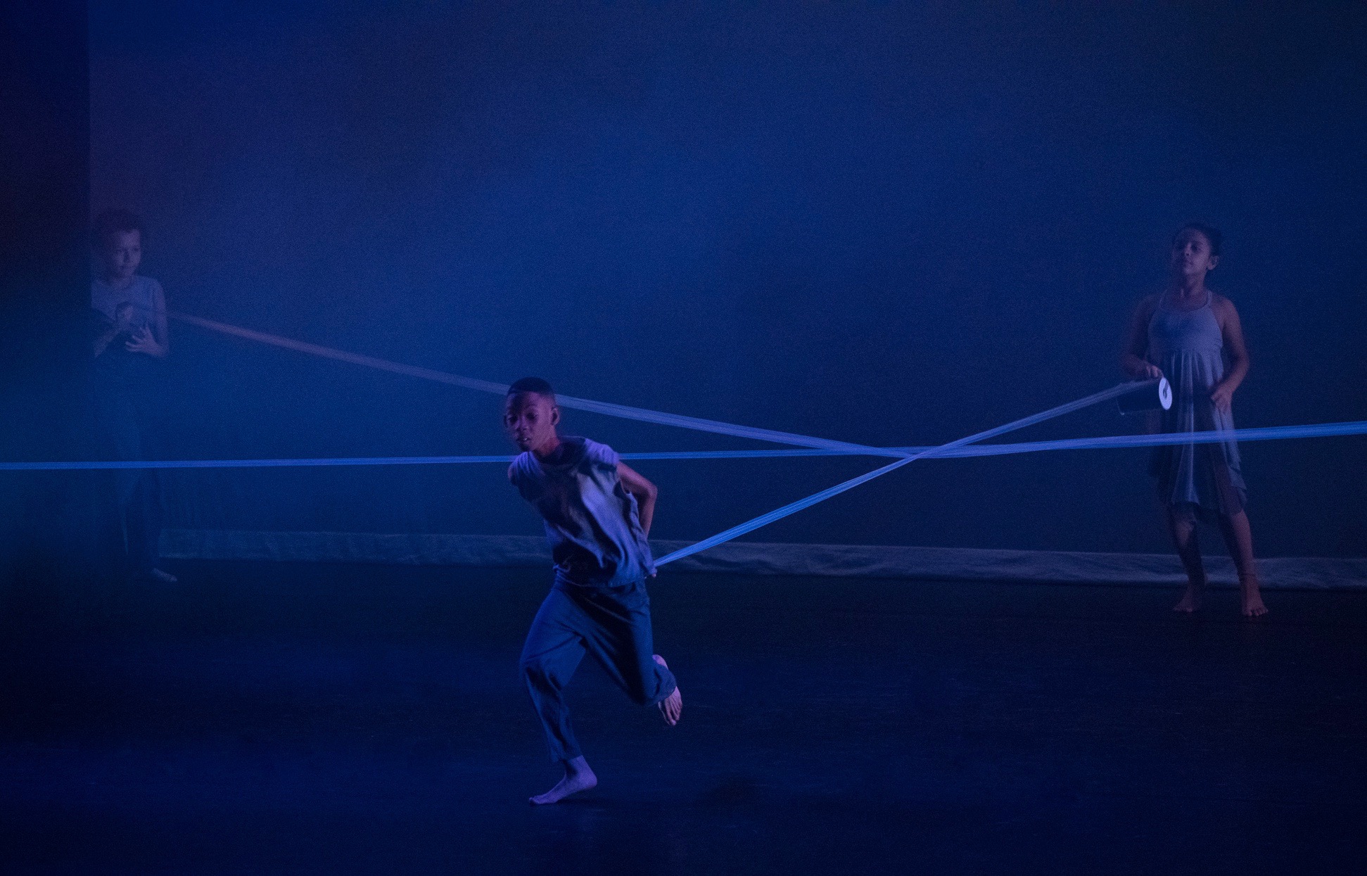 Grey. Three student-dancers stand in different corners of the stage holding longs strips of grey fabric that create a star-shaped web across the stage. The stage is lit in dark blue.