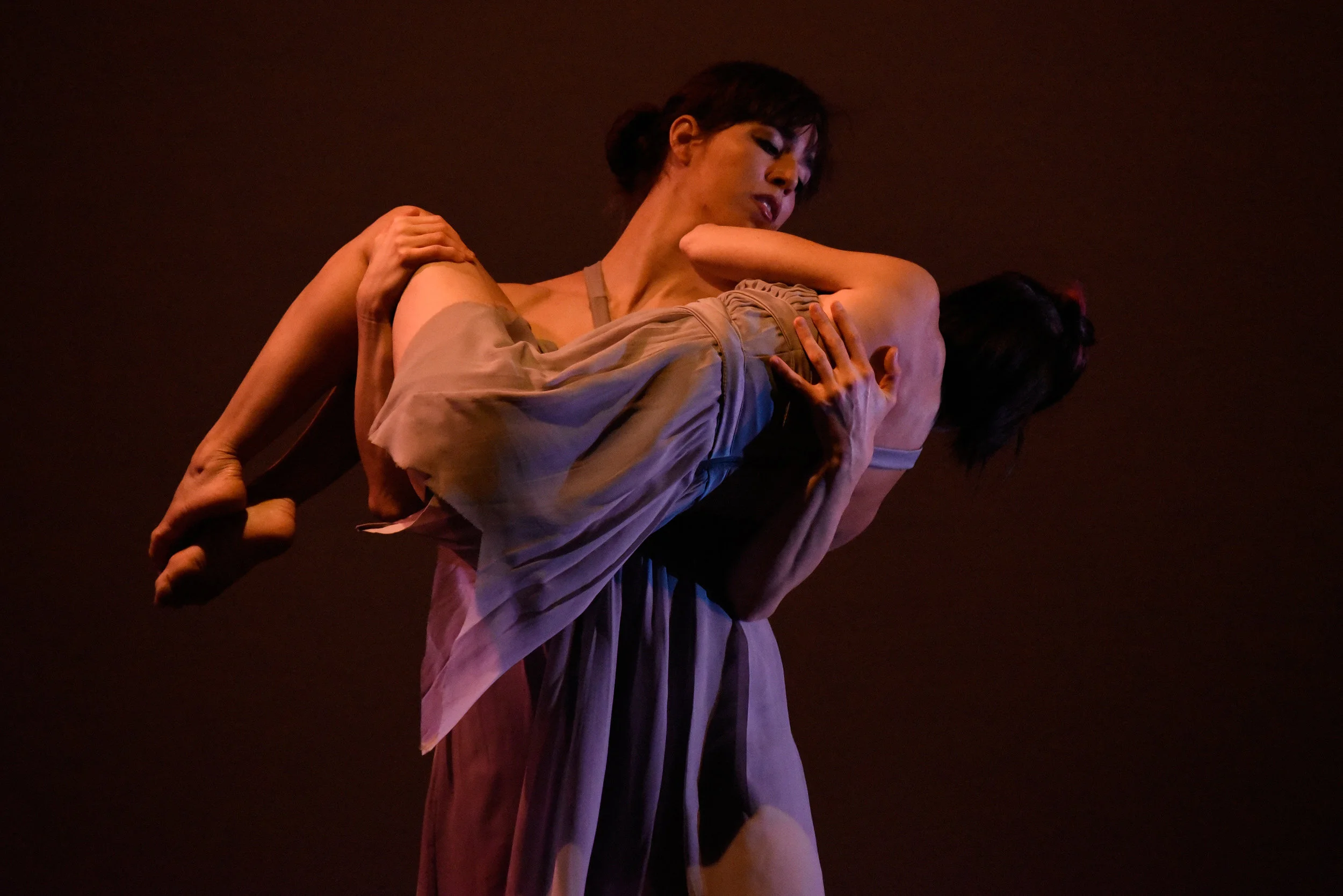 Grey. Dancer Kelly Vaghenas holds and carries dancer Lisa Clementi as Lisa is folded into a fetal position and wraps around Kelly’s chest.