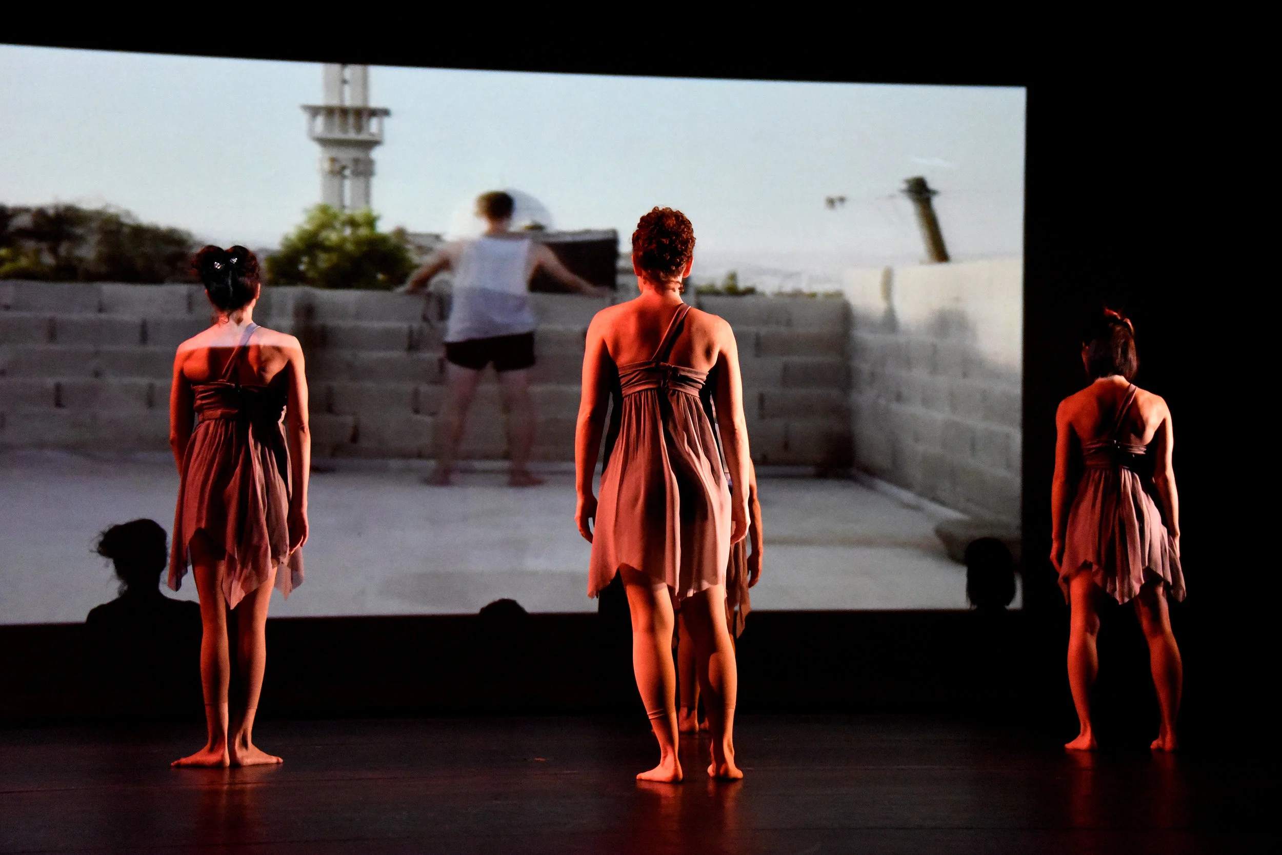 Grey. Three women in short grey dresses stand still and face the backdrop showing a video of a dancer dancing on a rooftop in Syria.