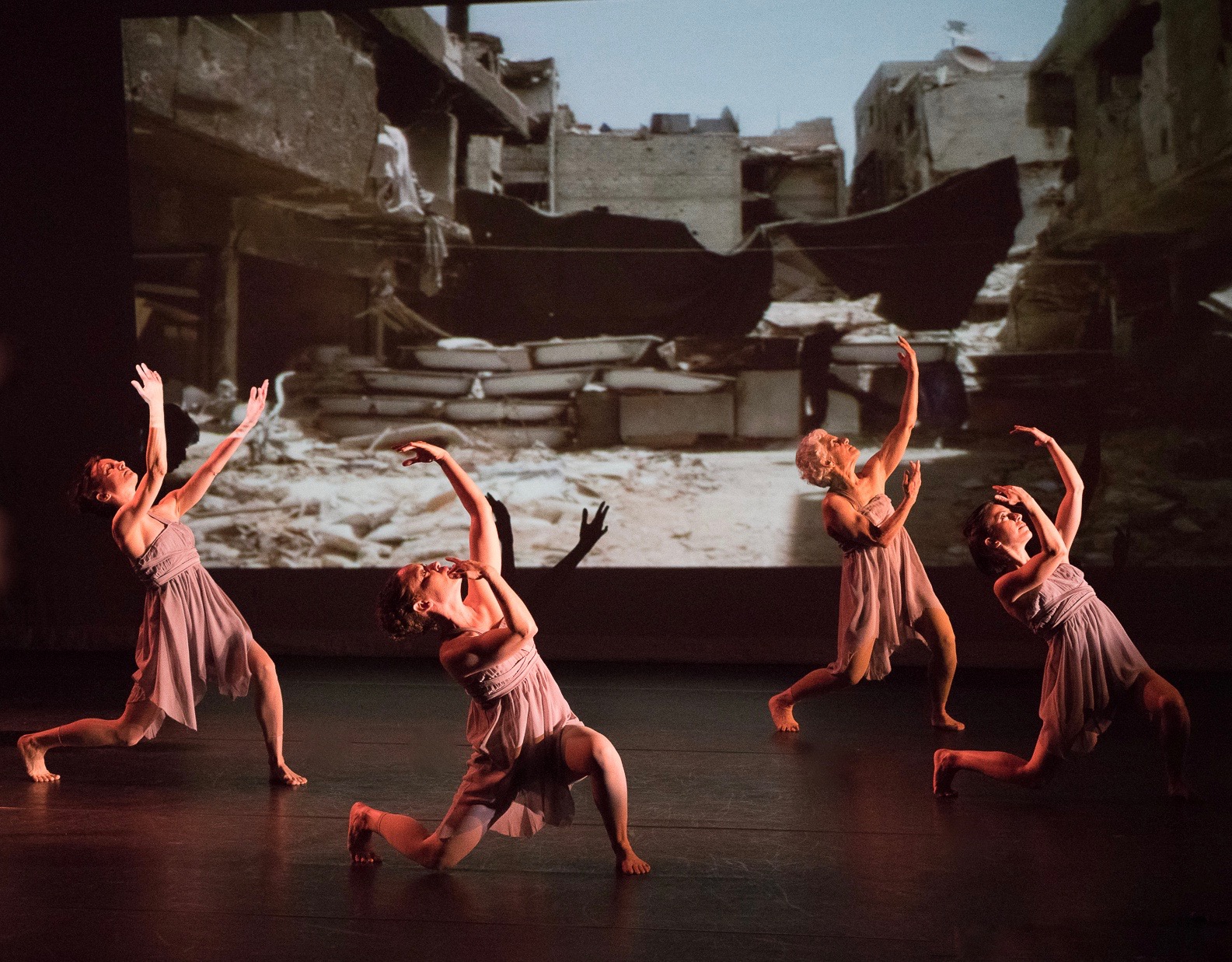 Grey. Four women bend their legs and arch their bodies back as they curve their arms and look up. Behind them on the backdrop is an image of a street in Syria that has been destroyed.
