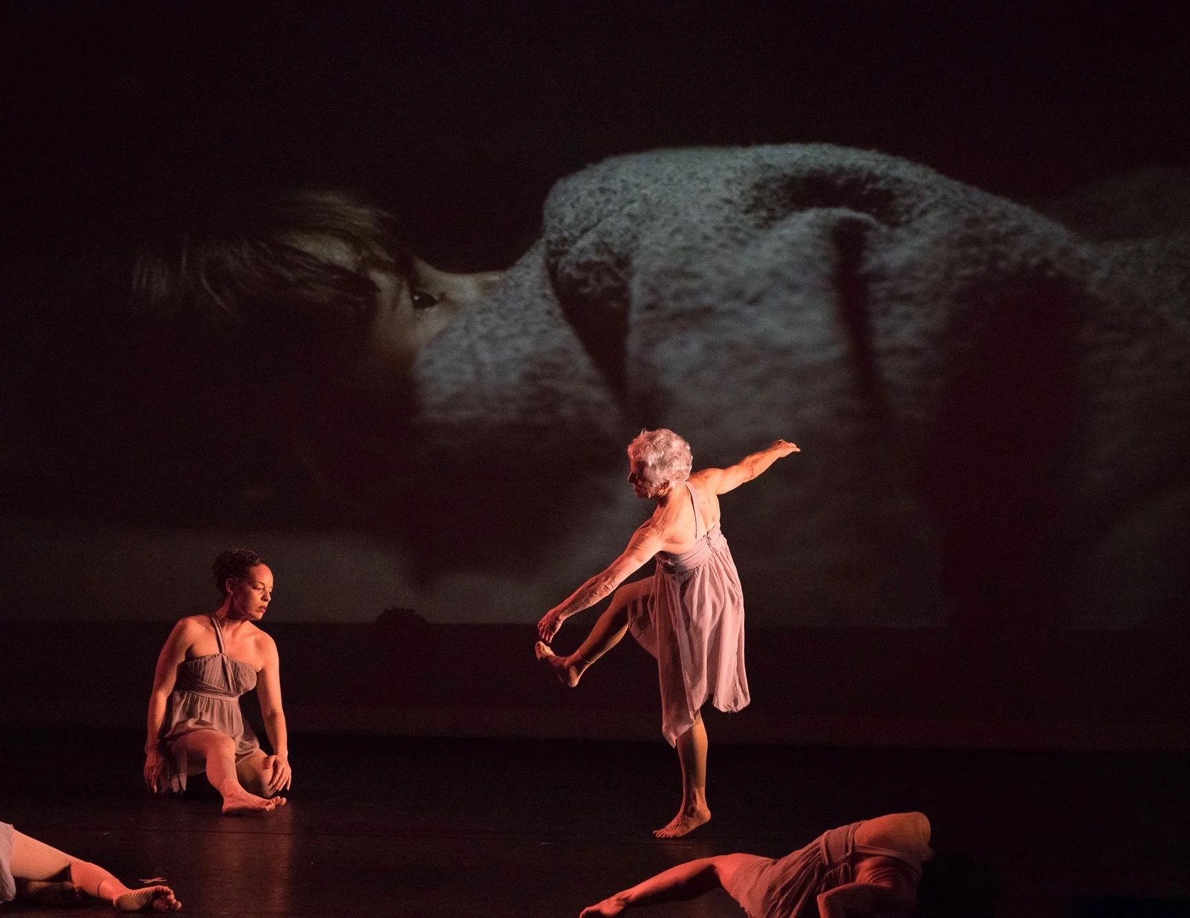 Grey. On a dimly lit stage dancer Janet Nightingale turns and lifts her leg in front of an image of Farah, who fled Syria and loves soccer. (Photo by Magnus Wennman ‘Where the Children Sleep’ exhibit. Photo used by permission)