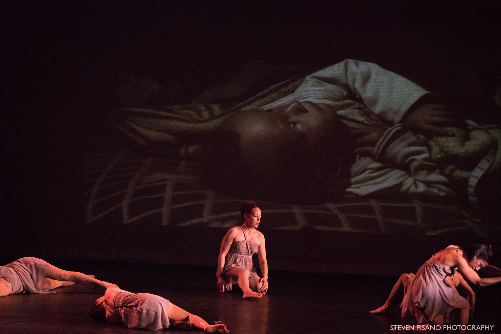 Grey. Dancers lay down and sit in front of a photo of toddler Amir looking up from his makeshift bed and who was born a refugee and doesn’t speak. (Photo by Magnus Wennman ‘Where the Children Sleep’ exhibit. Photo used by permission)