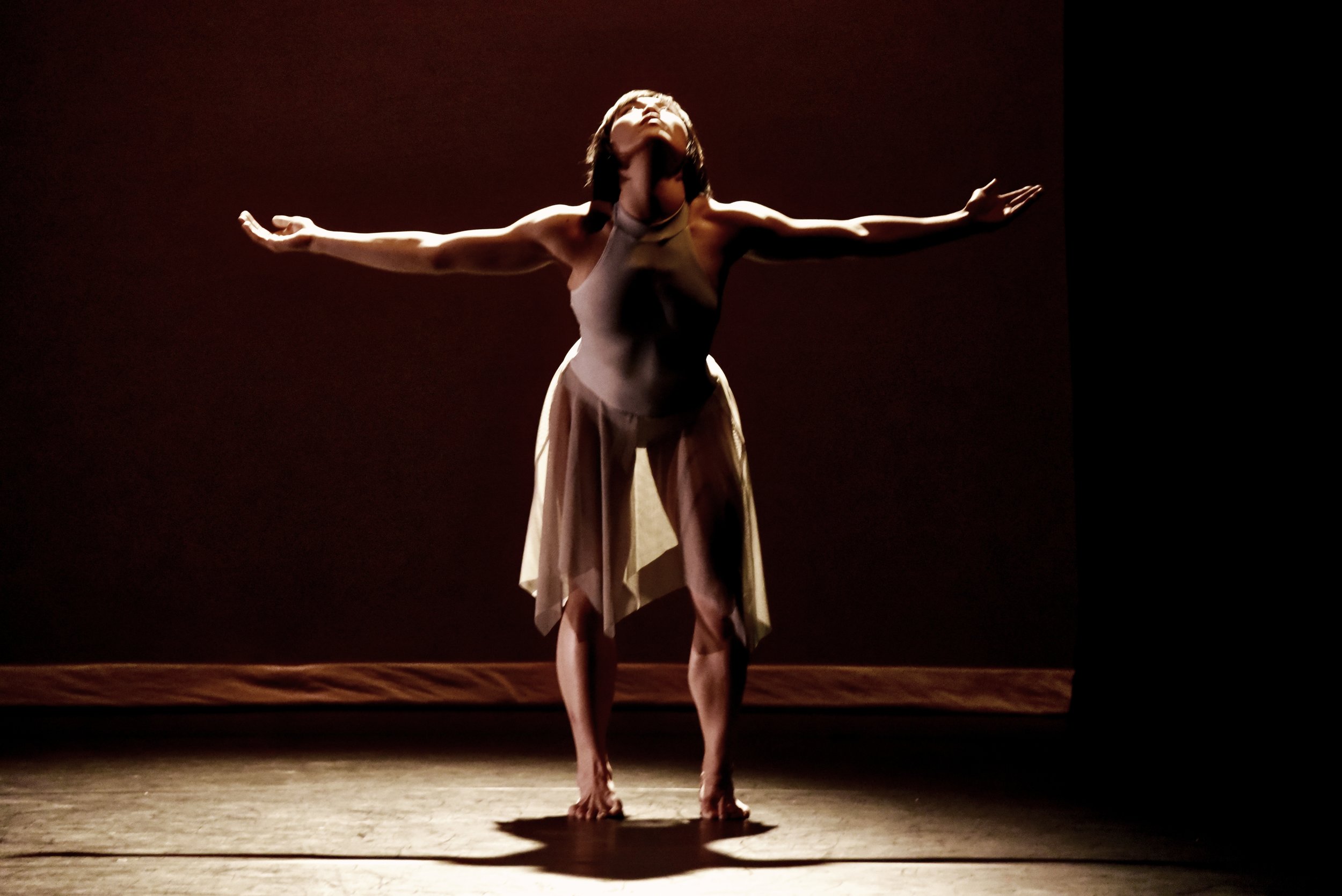 Grey. Dancer Tina Wang in a soft grey leotard and light skirt reaches both arms out to the side and arches her back and looks up.