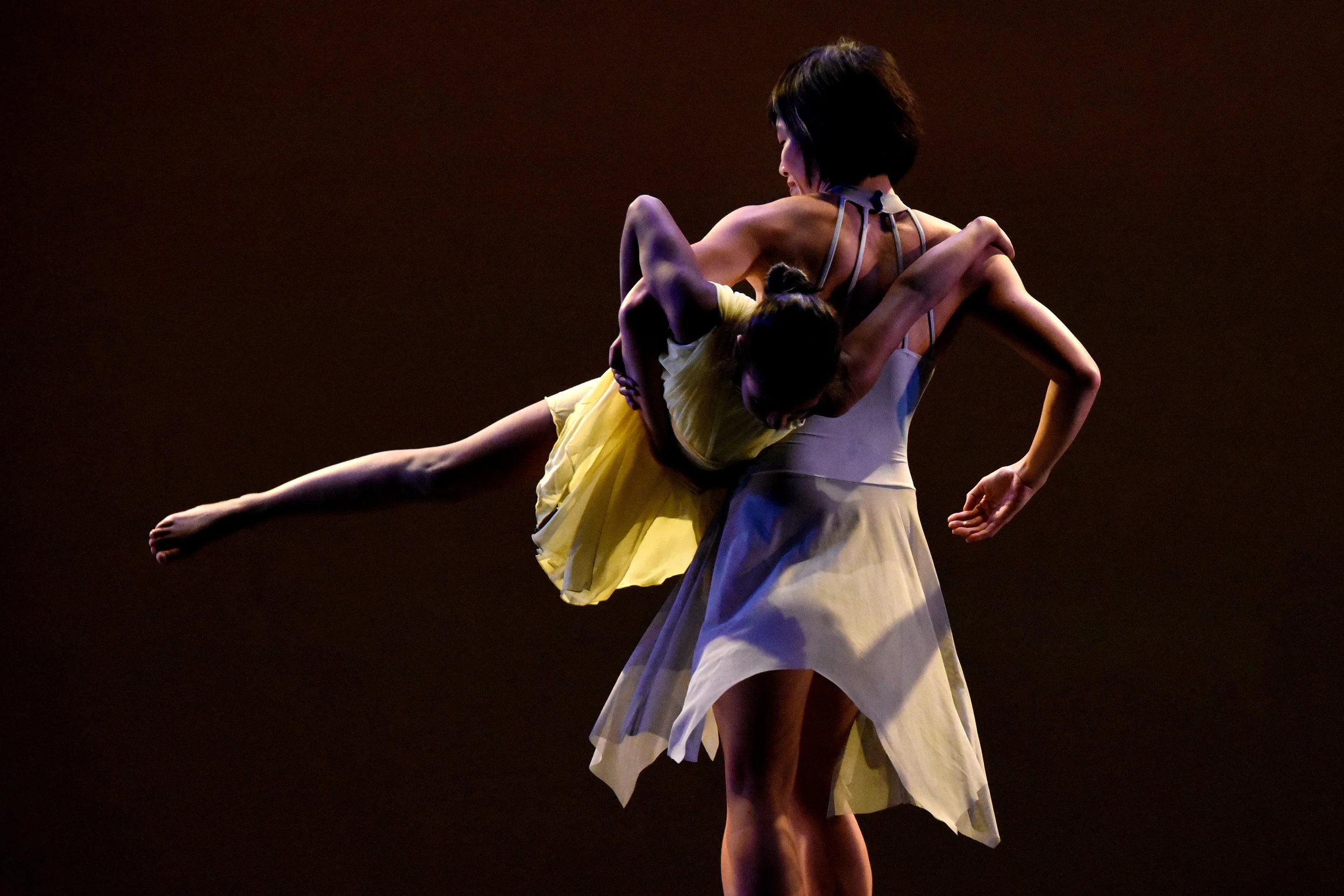 Grey. On a stage dimly lit in amber dancer Tina Wang lifts student-dancer Emily and swirls her around her trunk. Emily holds onto Tina with her arms and has her legs extended to the side as she swirls.