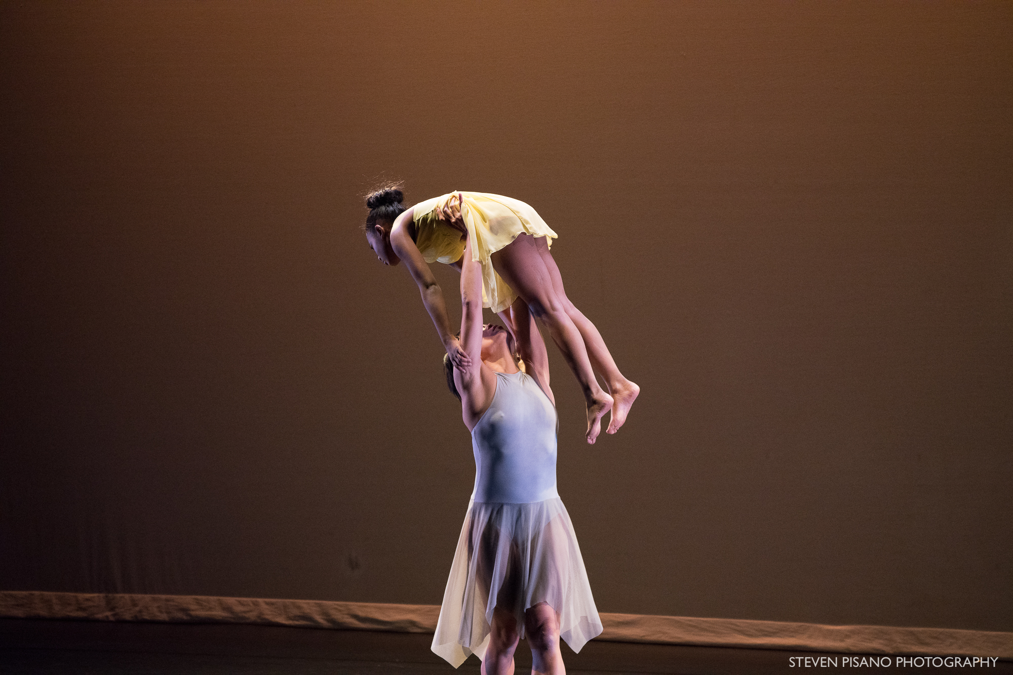 Grey. Against a soft grey and beige backdrop dancer Tina Wang lifts student-dancer Emily high above her head.