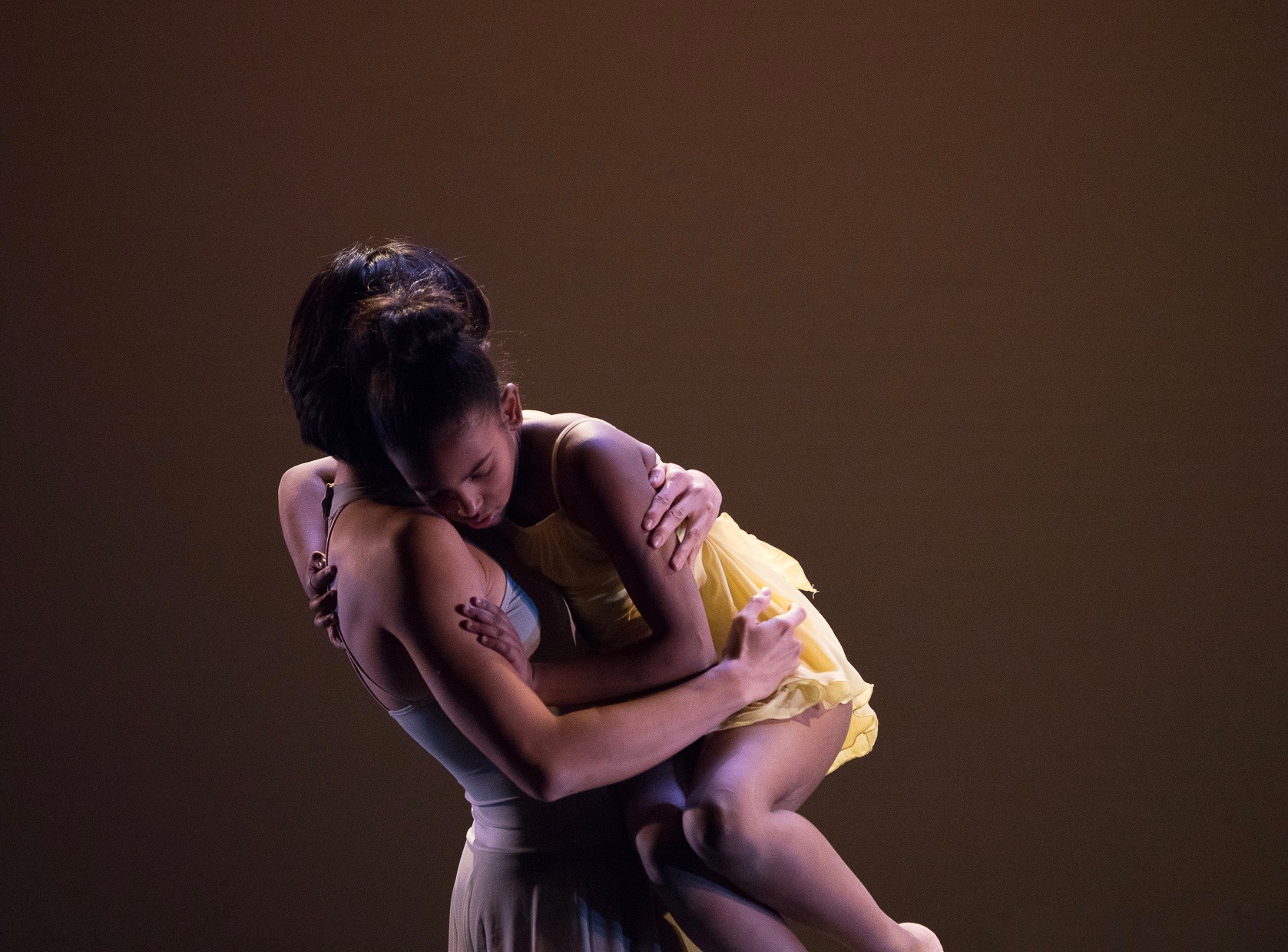 Grey. Against a soft grey and beige backdrop dancer Tina Wang holds student-dancer Emily folded against her chest as she carries her. Emily rests her head against Tina’s shoulder.