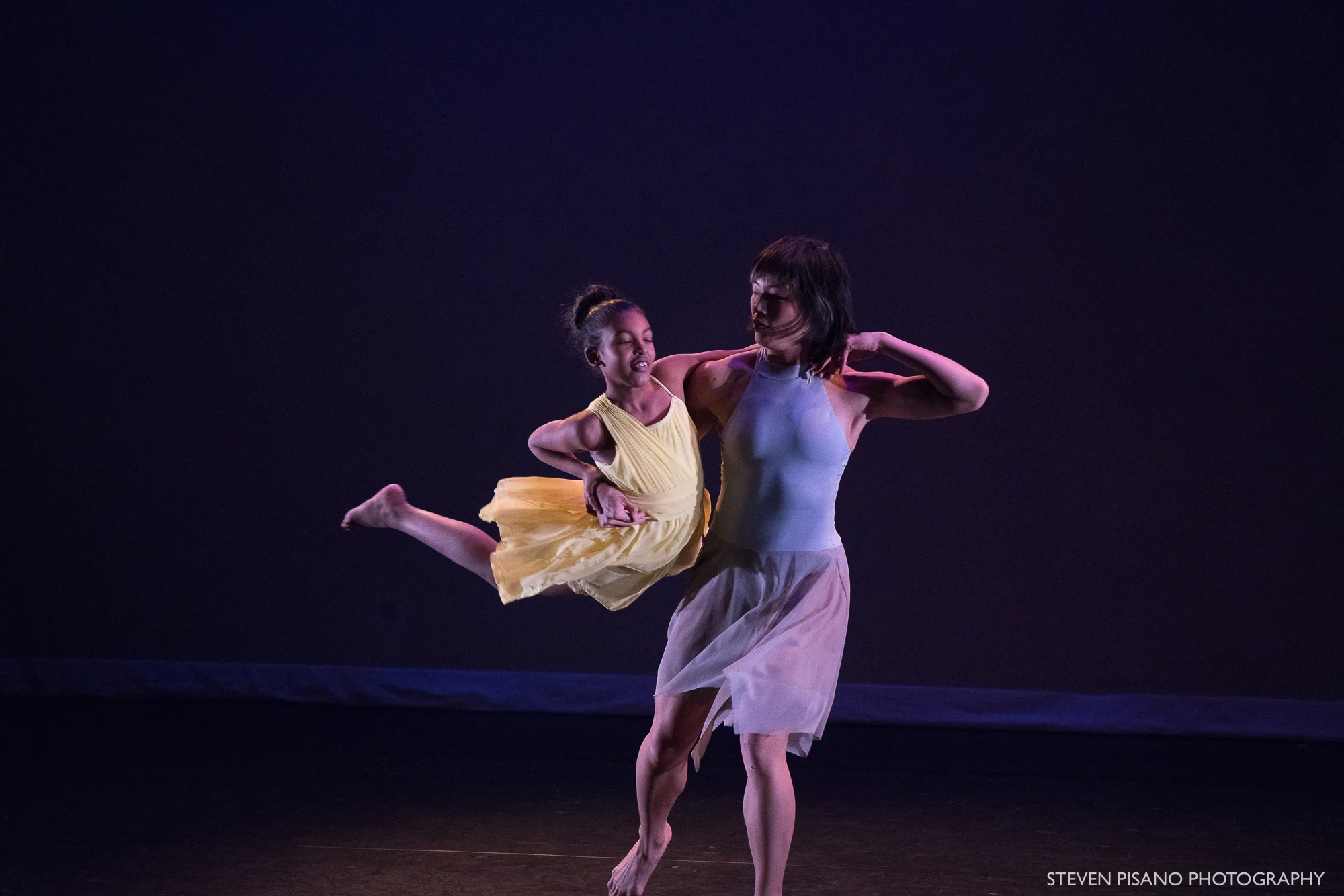Grey. Against a very dark blue backdrop dancer Tina Wang lifts student-dancer Emily and swirls her around her trunk. Emily holds onto Tina with her left arm and has her legs extend out behind her.