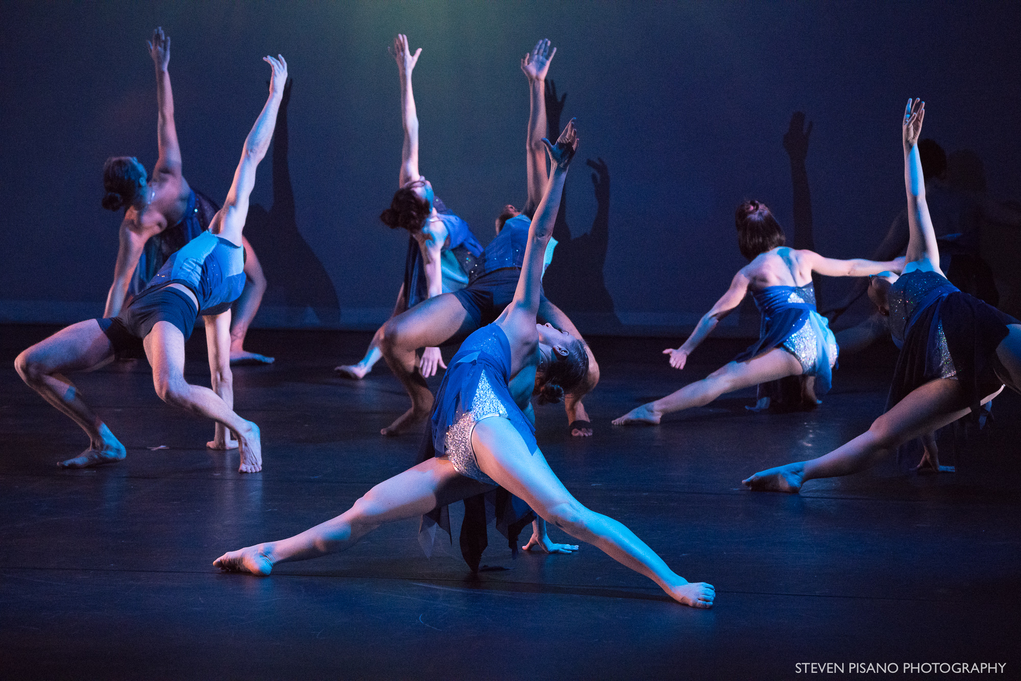 Silver Staircase. Against a backdrop that has an image projected of being under water, seven dancers are in a side push-up pushing off on one arm with the other arm reaching straight up as they reach out to the side with both legs straight and extended.