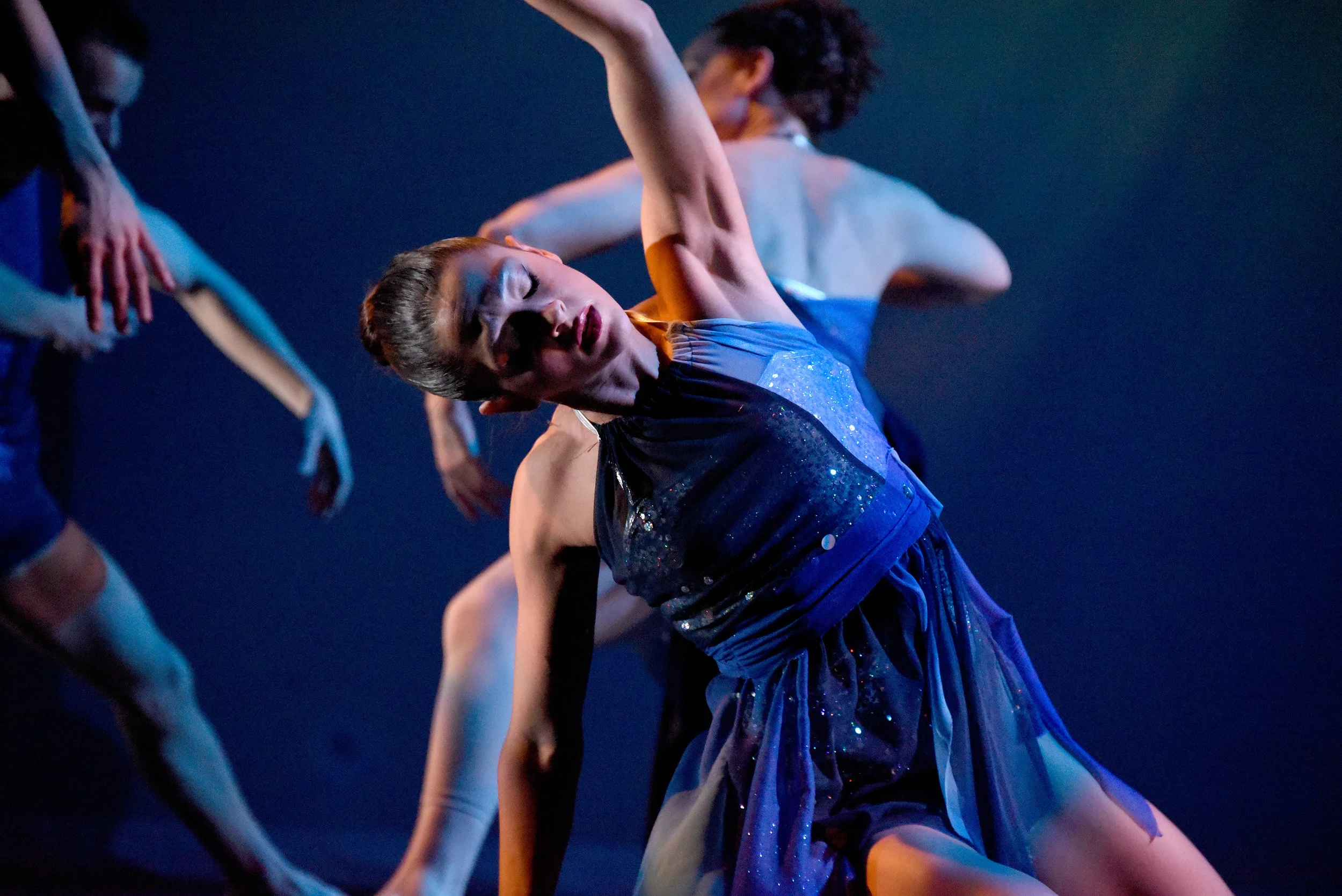 Silver Staircase. Against a backdrop that has an image projected of being under water, dancer Stefanie Roper in a short wavy dress in various colors blue and silver, sits on her knees and bends to one side as she reaches her arm overhead.