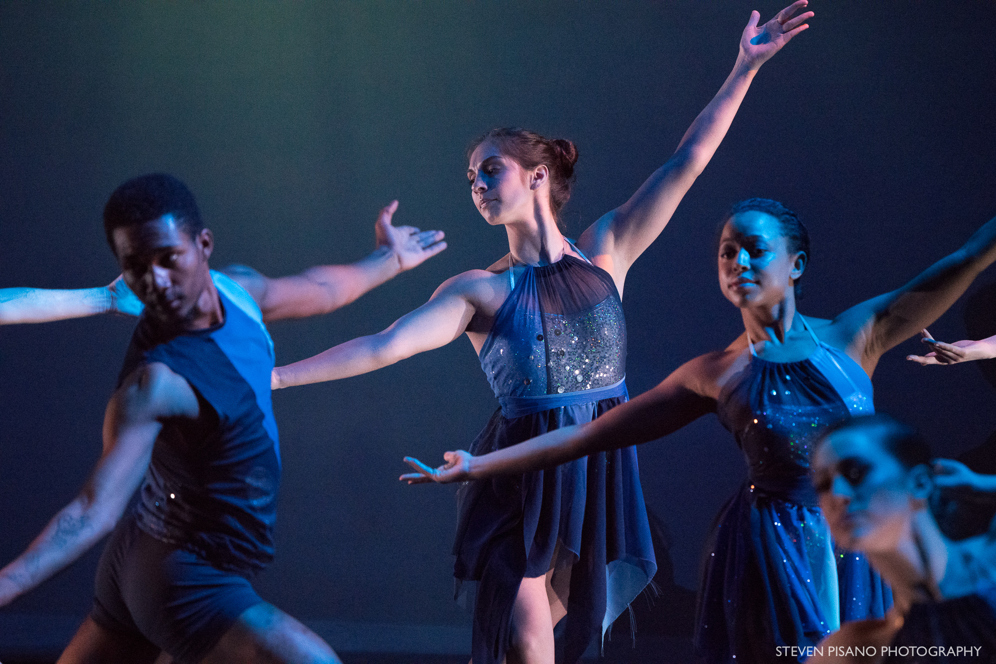 Silver Staircase. Close up of four dancers in dark blue and other shades of blue and silver wavy short costumes. All four dancers reach out with their arms in a large semi-circle shape.