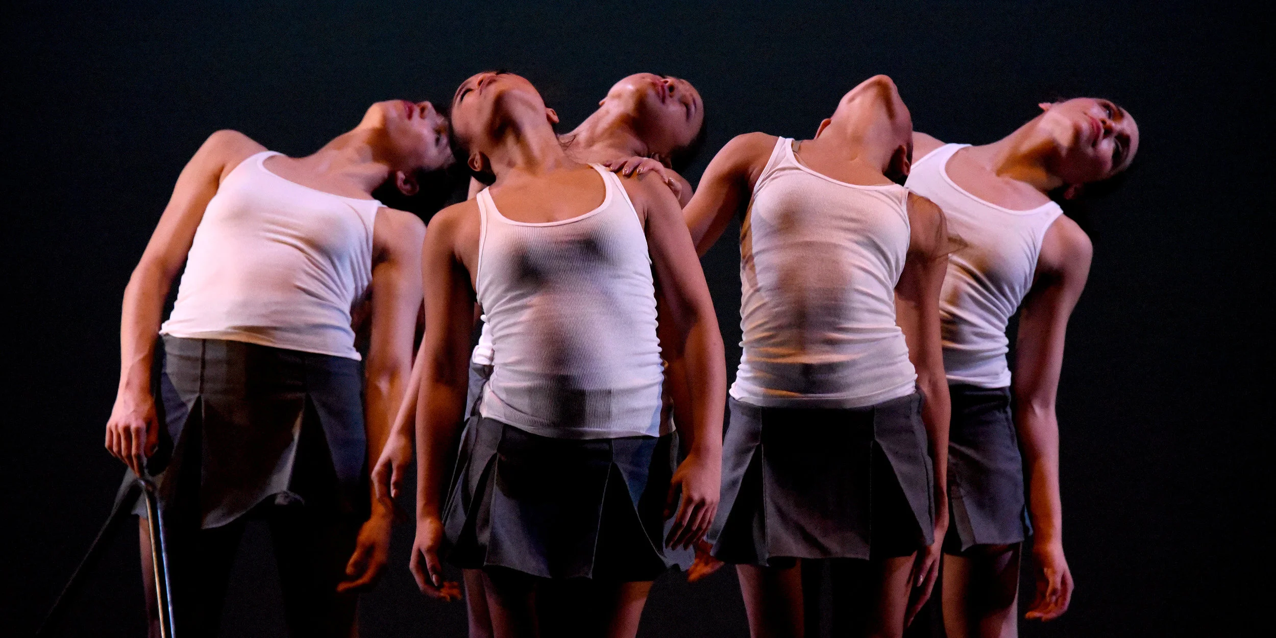 Sshhh. Five women dressed in grey school uniform skirts and white tank tops stand together as they arch their backs and rotate their upper bodies.