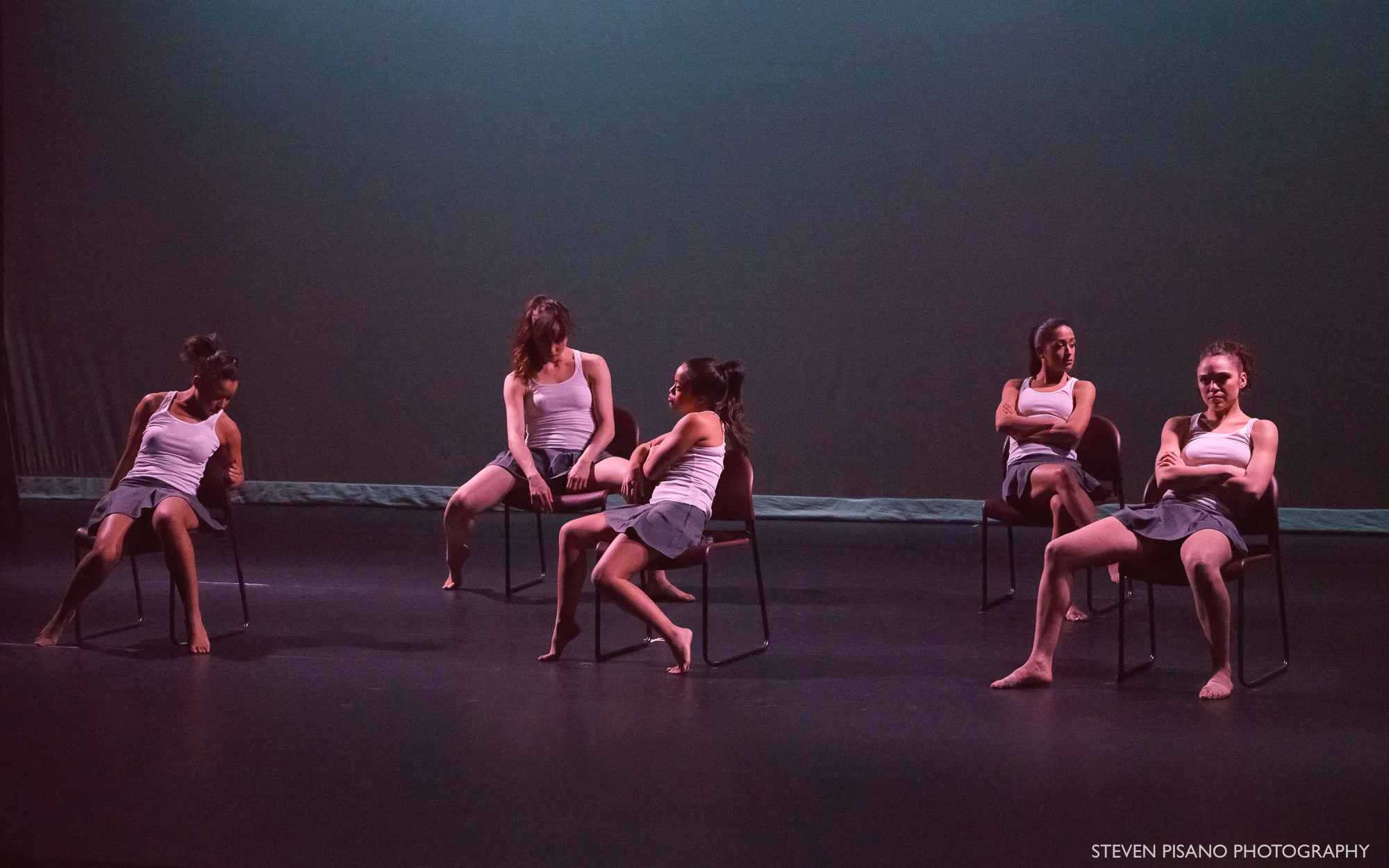 Sshhh. Five women dressed in grey school uniform skirts and white tank tops sit on five chairs in various slouched positions and they look like they are waiting or bored. The stage is lit in a dark teal and grey color.