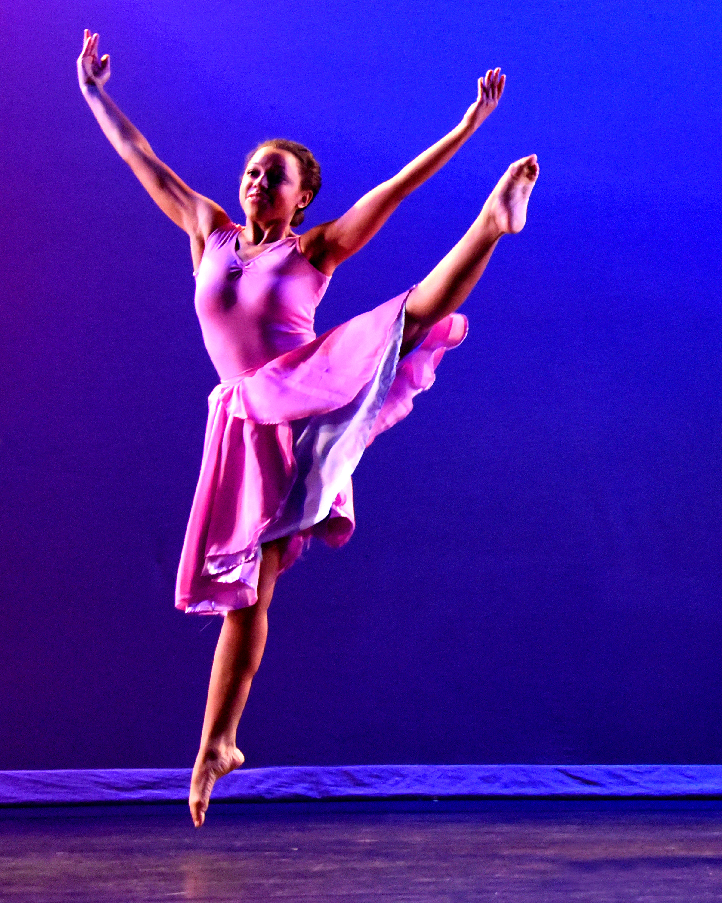 Stems. Dancer Amber Davis jumps with one leg kicked up high to the side and both arms up in a large V-shape against a blue and violet backdrop.