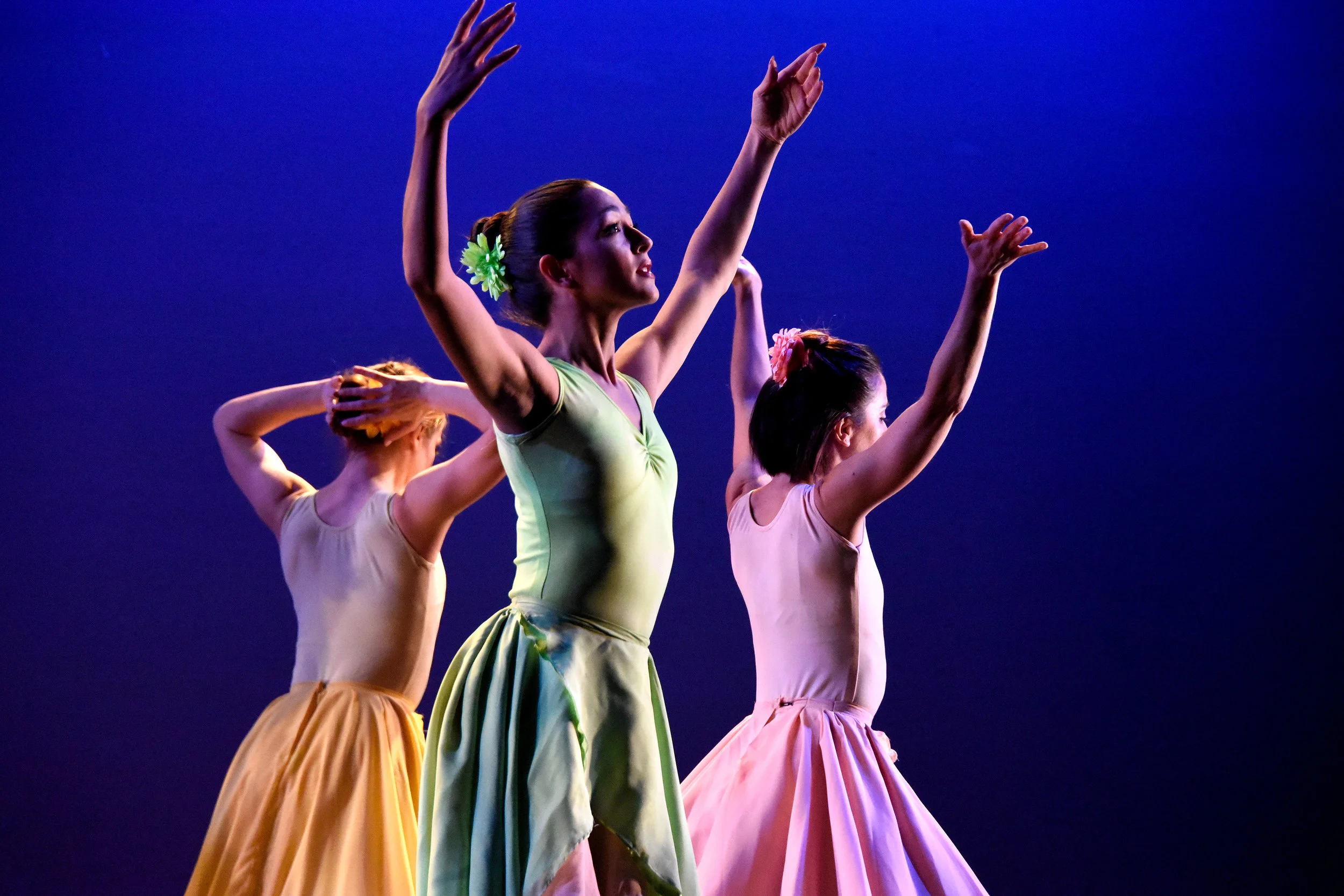 Stems. Close up of dancer Stefanie Roper in a light green dress reaching up with both arms reaching out. Behind her are two women in the same pose in a yellow dress and a peach-colored dress.