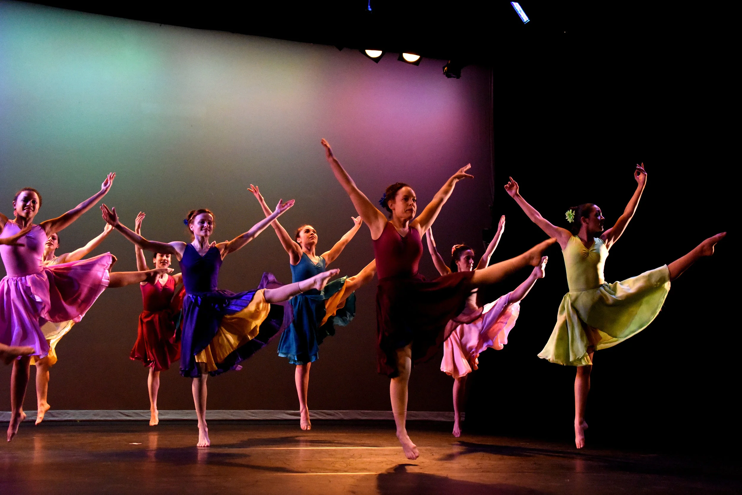Stems. Eight women in eight bright colored flowing knee-length dresses jump in unison with one leg kicked up high to the side and their arms reaching up in a large V-shape. The backdrop has a mix of pink, yellow, and teal colors.