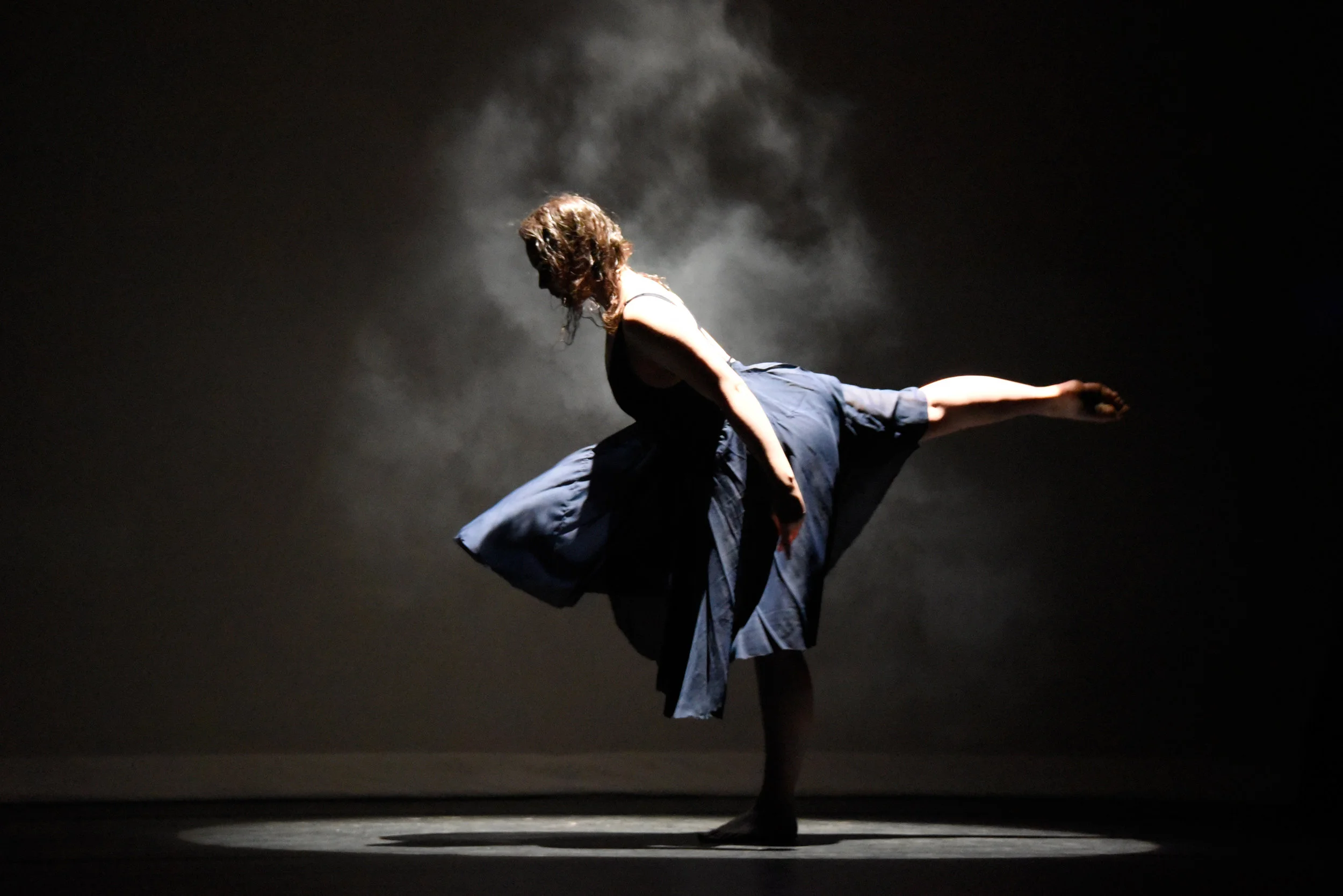 Stems. Dancer Katie Mollison in a dark blue knee-length flowing dress stands facing the side in a white foggy spotlight on a dark stage as she leans forward and reaches one leg behind her in arabesque.