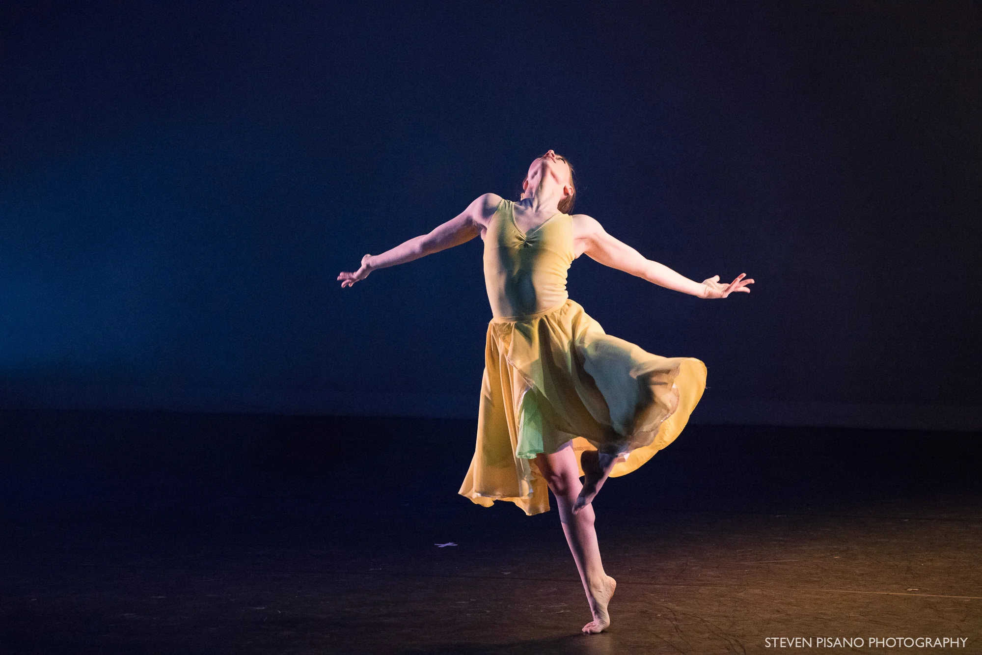 Stems. Dancer Madeline Charles in a flowing daffodil-yellow dress stands on leg on her toes with the other leg bent at the knee creating a triangle shape while she arches her upper back and has her arms out to the side.