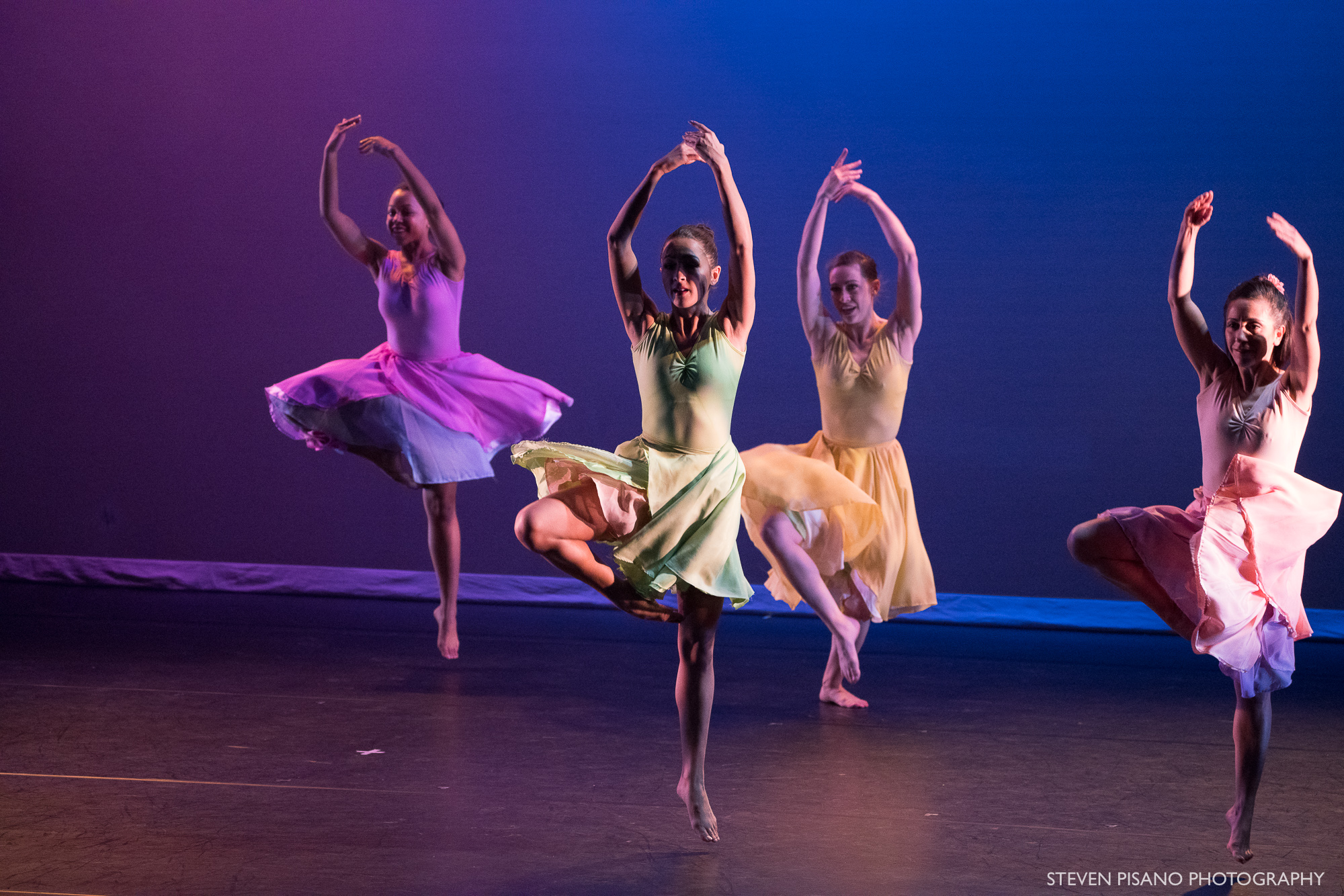 Stems. Four women in violet, light green, yellow, and peach colored flowing knee-length dresses jump in unison with one leg pulled up and bent at the knee and their arms reaching up.. The backdrop has a mix of pink and blue colors.